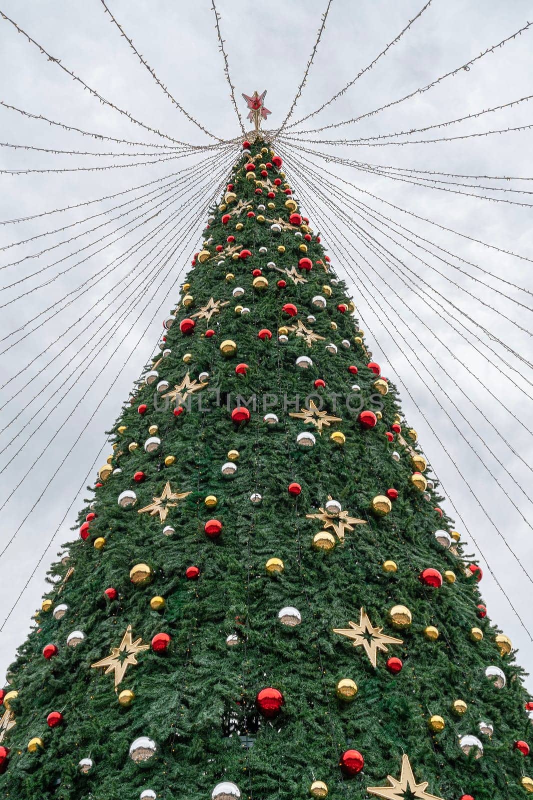 A festive Christmas tree towers high against a gray sky, decorated with colorful ornaments and shining lights, bringing holiday cheer to the outdoor setting.