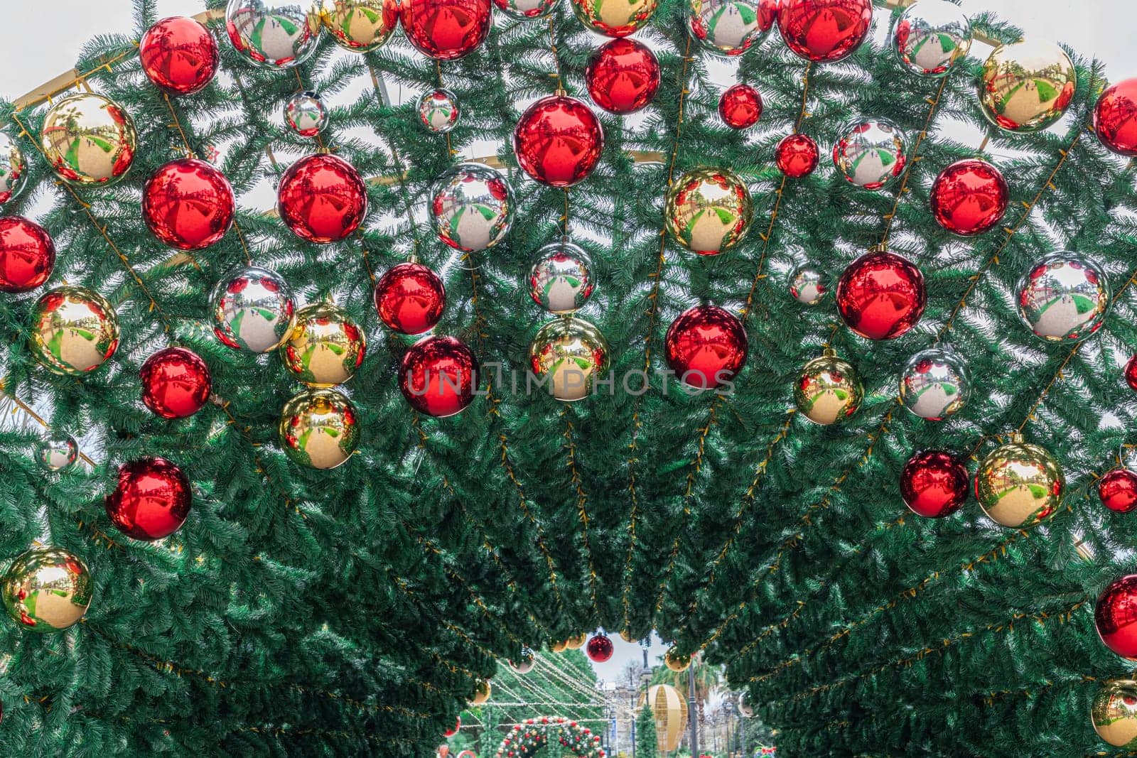 A beautifully adorned archway features vibrant red and gold ornaments amidst lush greenery, inviting visitors to explore a lively winter market filled with holiday spirit.
