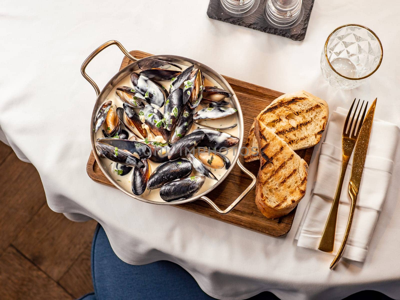 Delicious black mussels in creamy cheese and garlic sauce. Mediterranean black mussels in metal pan on wooden cutting board with grilled bread on restaurant table background, top view