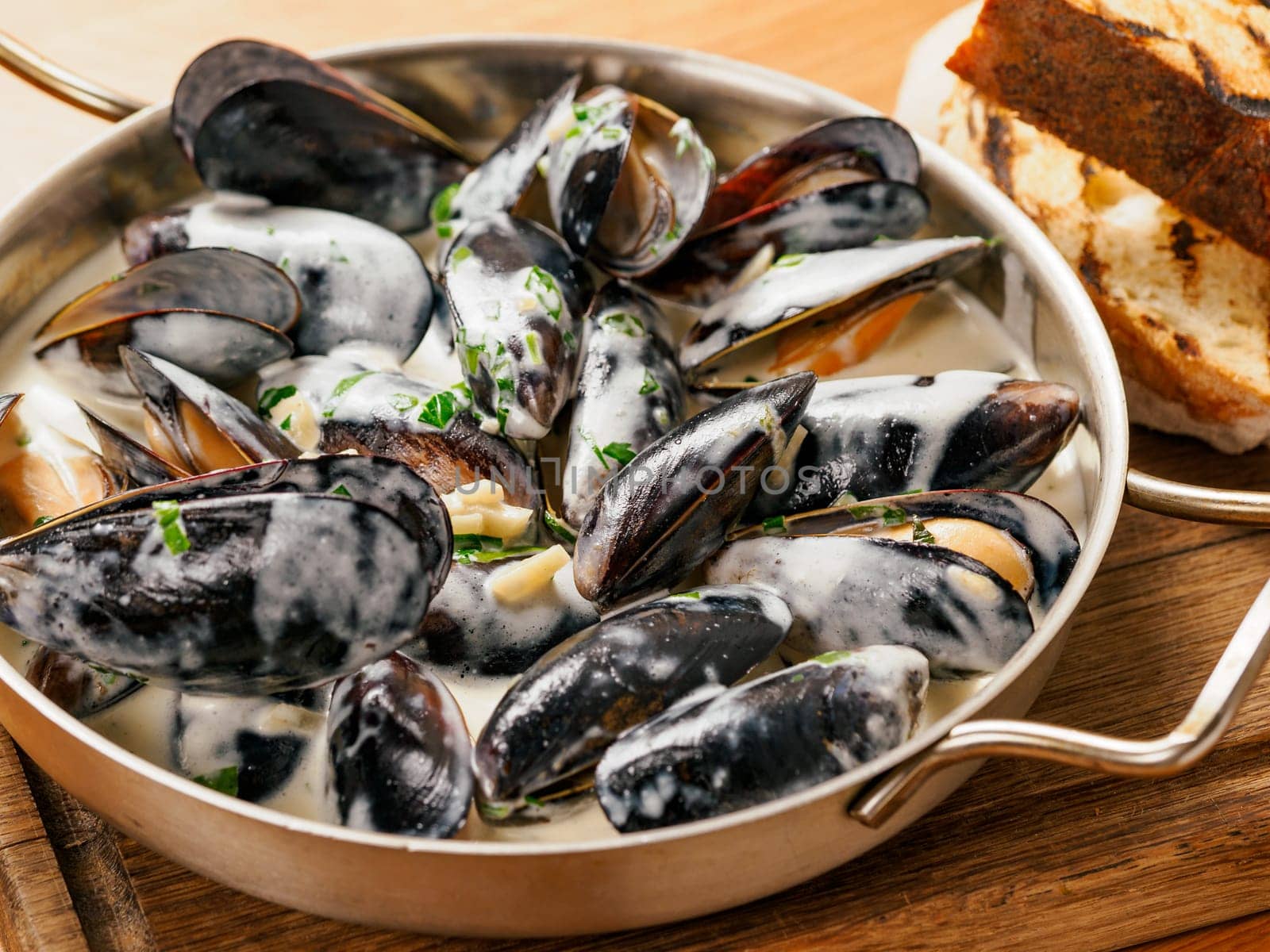 Delicious black mussels in creamy cheese and garlic sauce. Mediterranean black mussels in metal pan on wooden cutting board with grilled bread on wooden background
