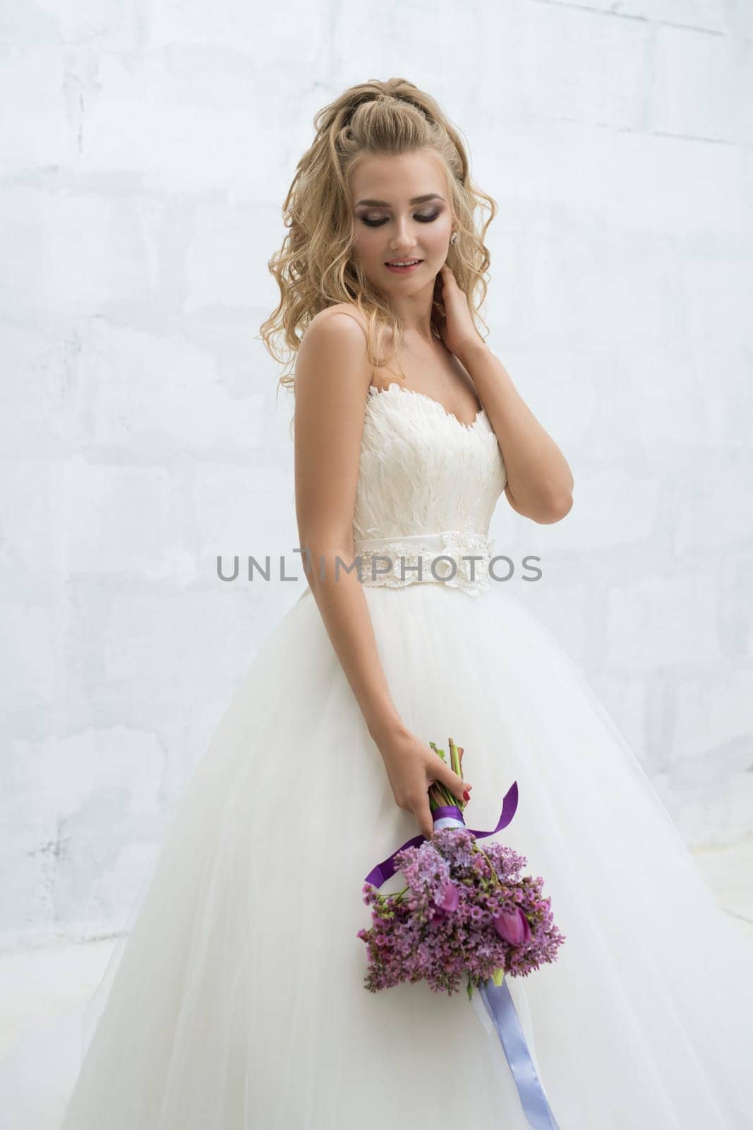 Gorgeous young bride in an exquisite dress posing with a violet bouquet in studio