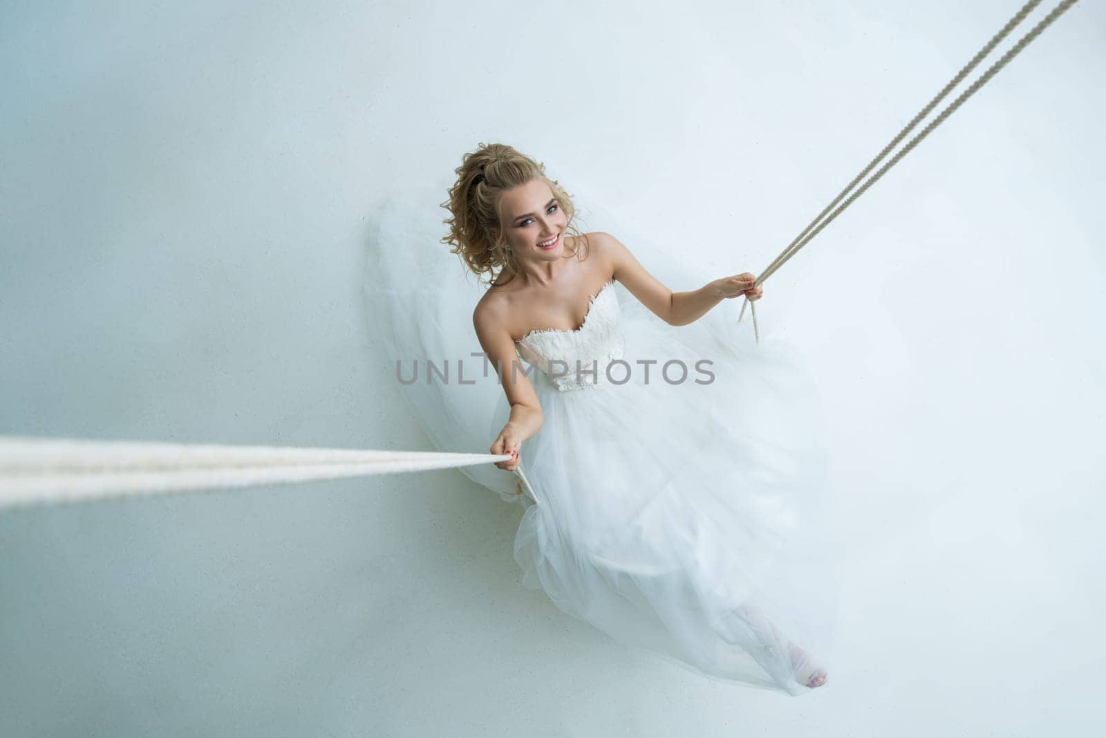 Gorgeous young bride in an exquisite dress posing on swing in white studio shot from above