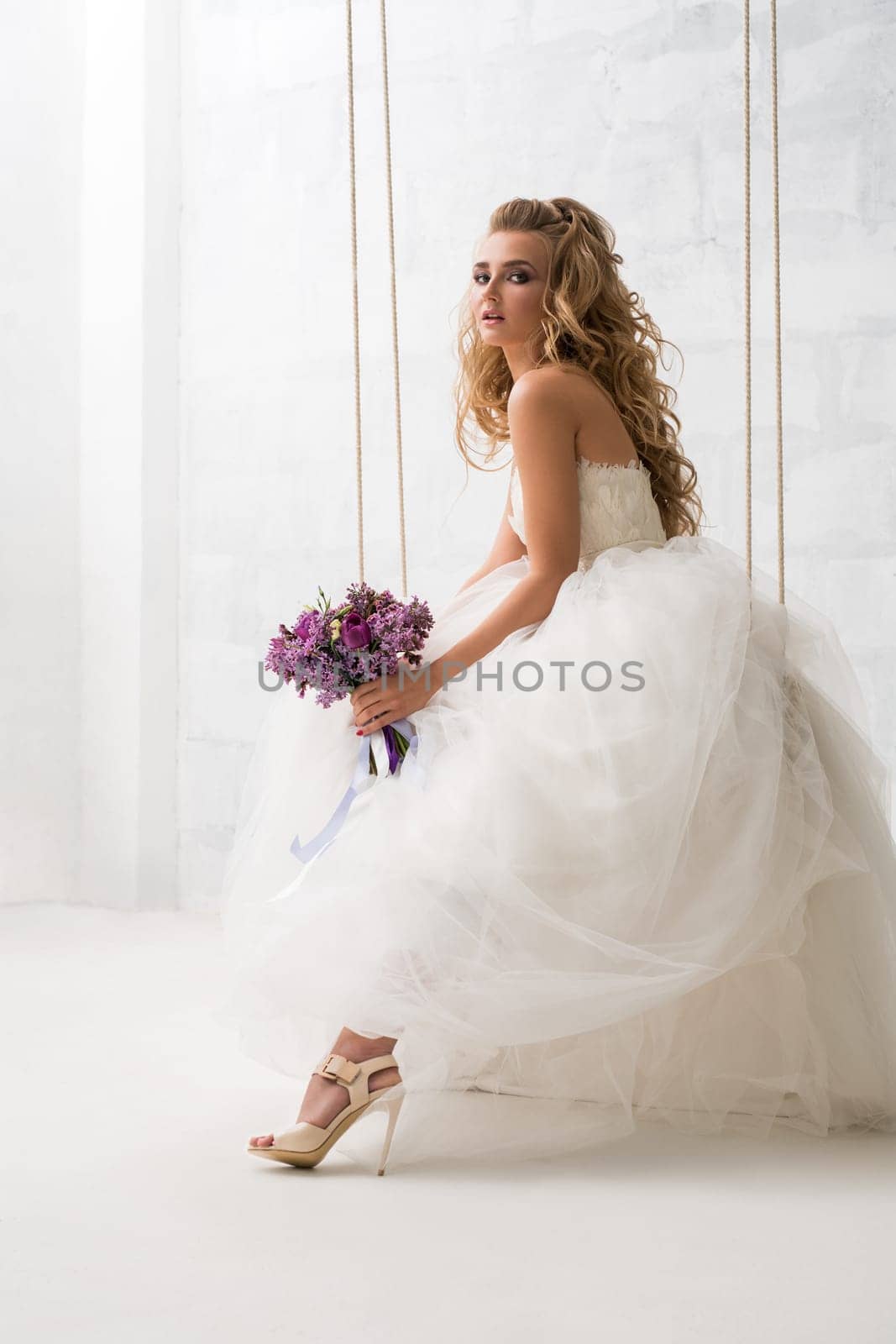 Young beautiful bride with gorgeous hair style posing on a swing holding nice bouquet studio shot