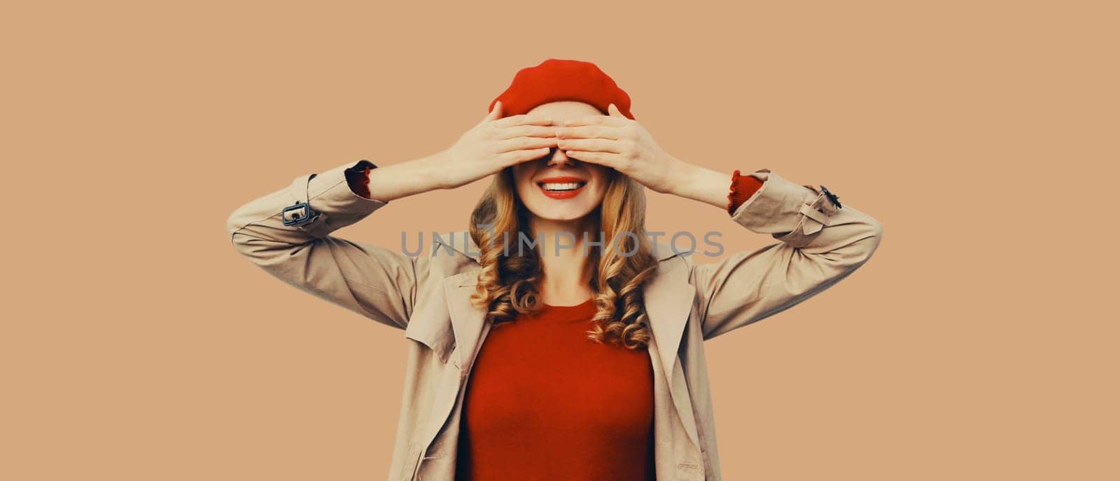 Stylish surprised young woman covering her eyes with hands, fashionable girl in red french beret hat, jacket posing on brown background