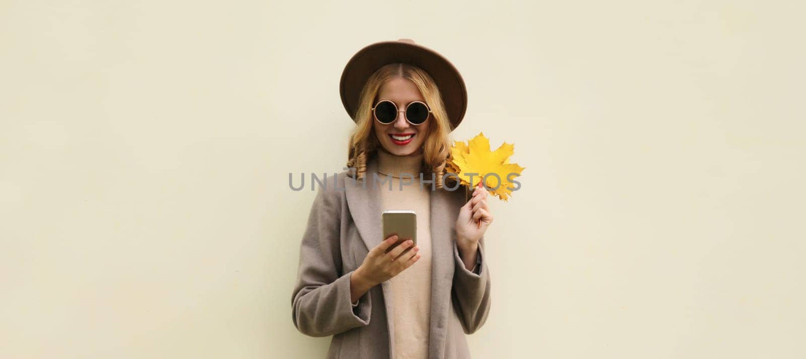 Autumn portrait of stylish happy smiling young woman with phone, beautiful girl holding yellow maple leaves in hat on white background