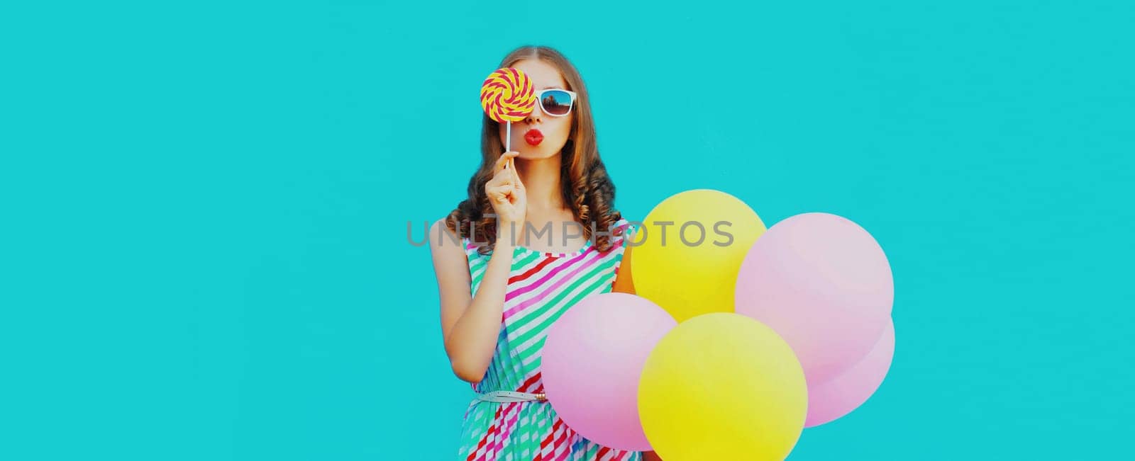 Beautiful young woman with bunch of balloons blowing kiss in colorful dress on blue background