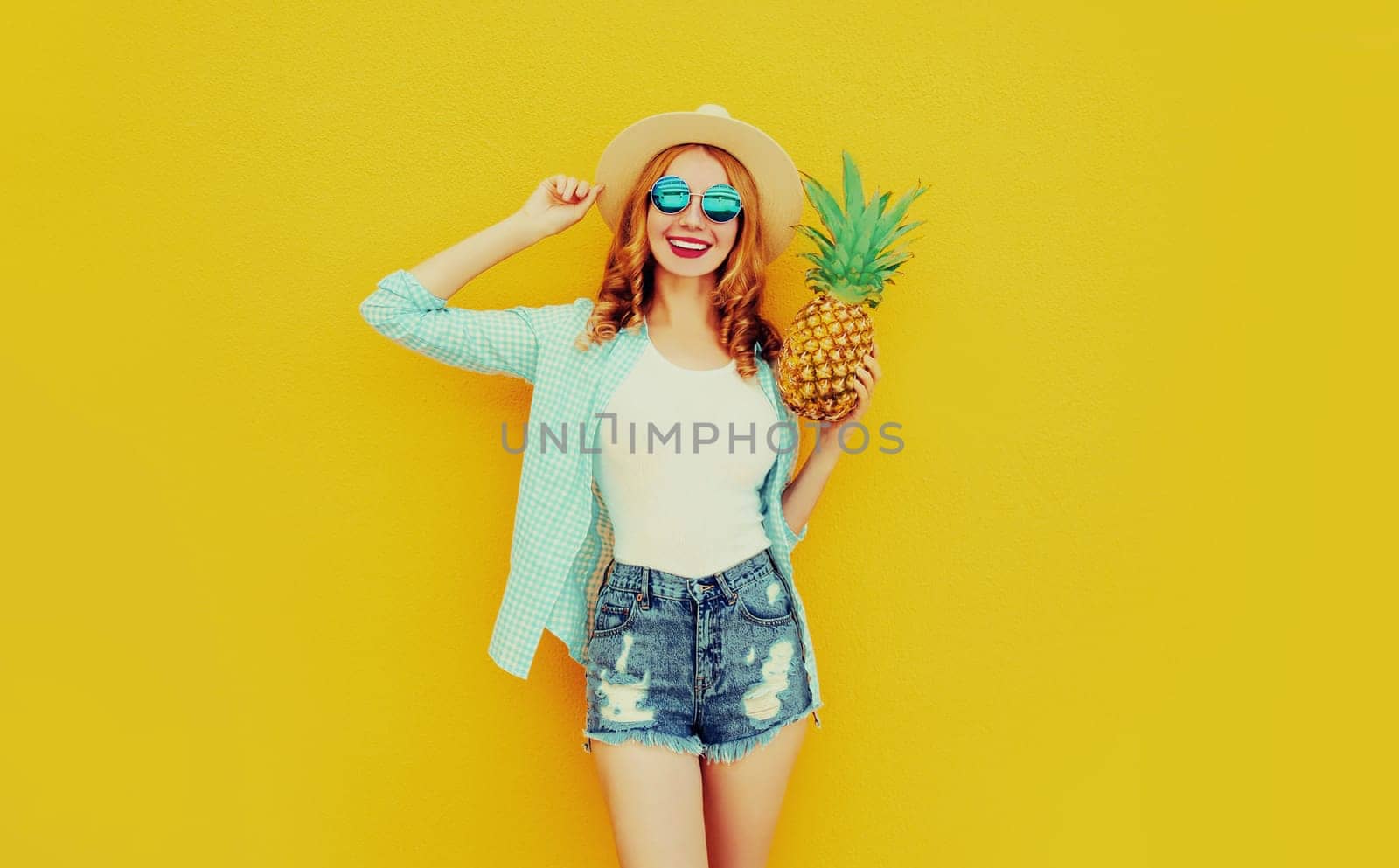 Happy smiling woman with pineapple having fun in summer hat, shorts on colorful yellow background