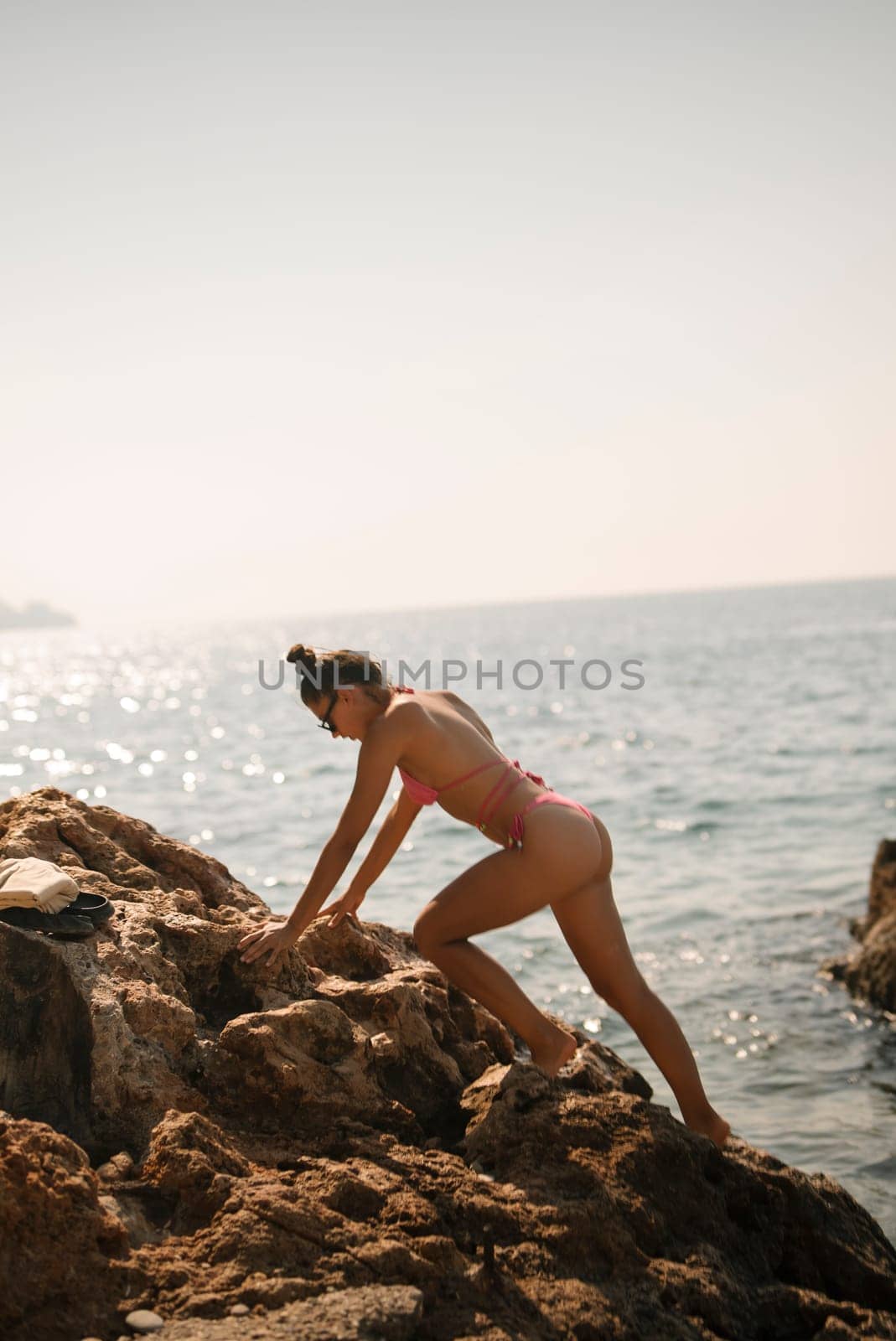 A woman climbs the rocky shores, delighting in the joys of summer by the shimmering ocean in a vibrant pink swimsuit