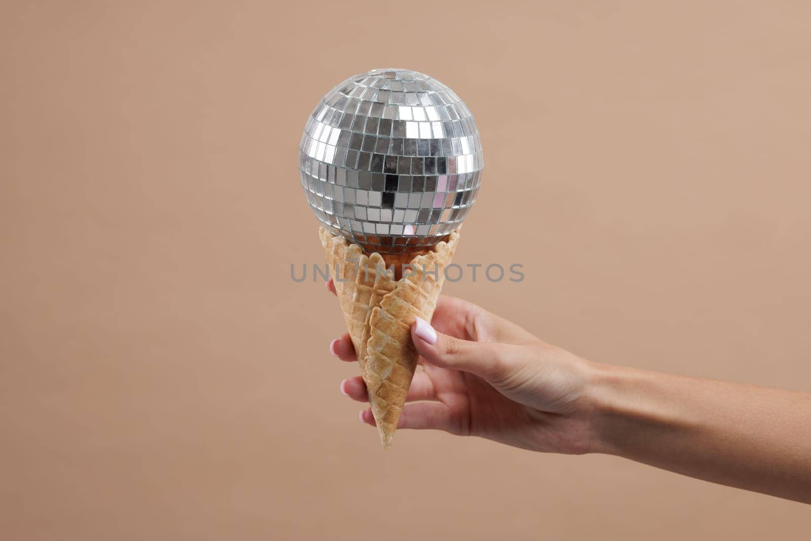 Hand holding waffle cone with disco ball on top, on neutral beige background. Disco party concept.
