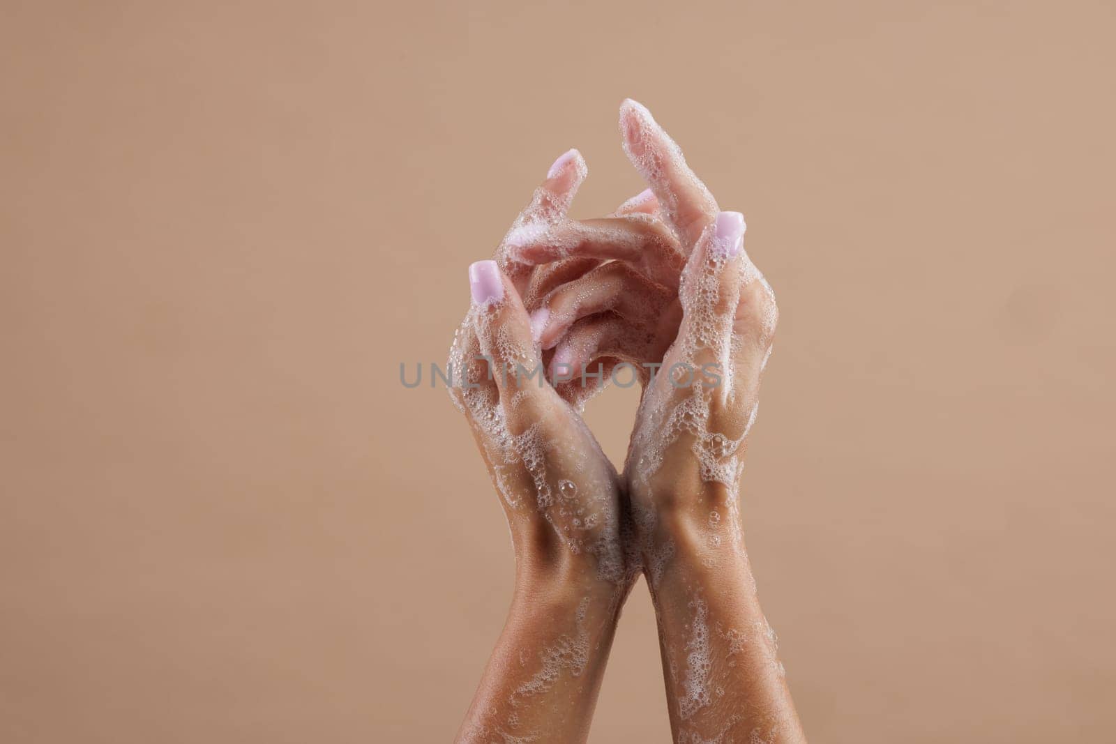A close-up of two hands lathered with soap, demonstrating hand washing. The hands are positioned in a way that emphasizes the soap bubbles against a neutral brown background.