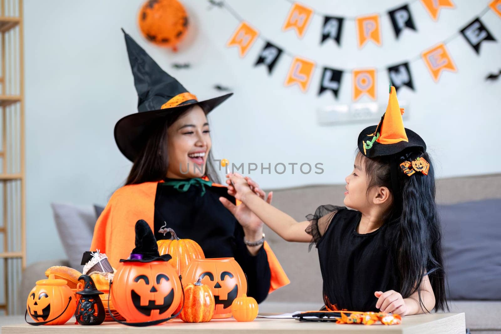 Playful young mother sitting on comfy couch in living room with cute little daughter tells spooky Halloween stories. Funny mom and girl kid have fun at home. Celebration and Autumn holiday concept.