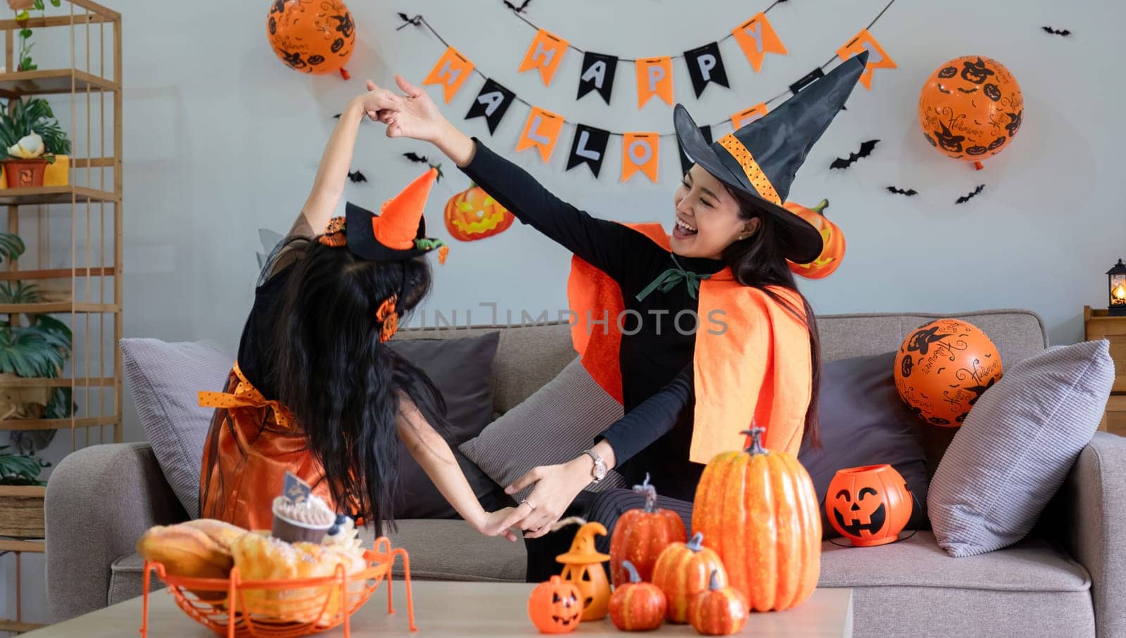 Playful young mother sitting on comfy couch in living room with cute little daughter tells spooky Halloween stories. Funny mom and girl kid have fun at home. Celebration and Autumn holiday concept.