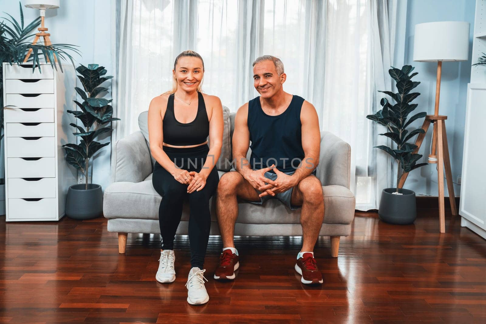 Athletic and sporty senior couple portrait in sportswear sitting on sofa as home exercise concept. Healthy fit body lifestyle after retirement. Clout