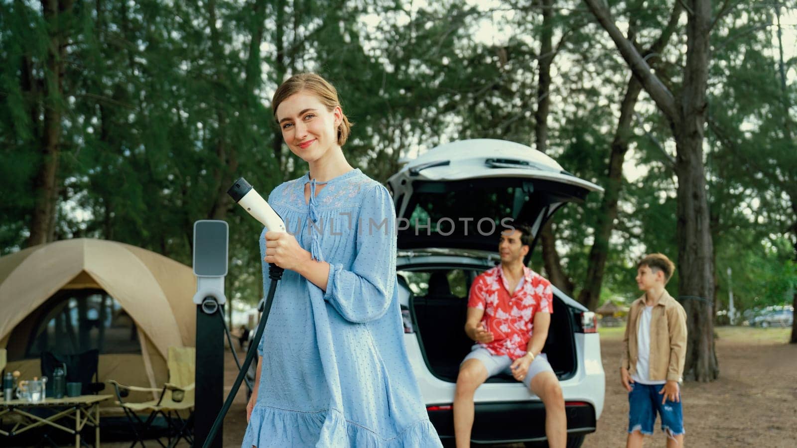 Outdoor adventure and family vacation camping at sea travel by eco friendly car. Cheerful woman or mother holding, pointing EV charger point with playful and happiness posture in campsite. Perpetual