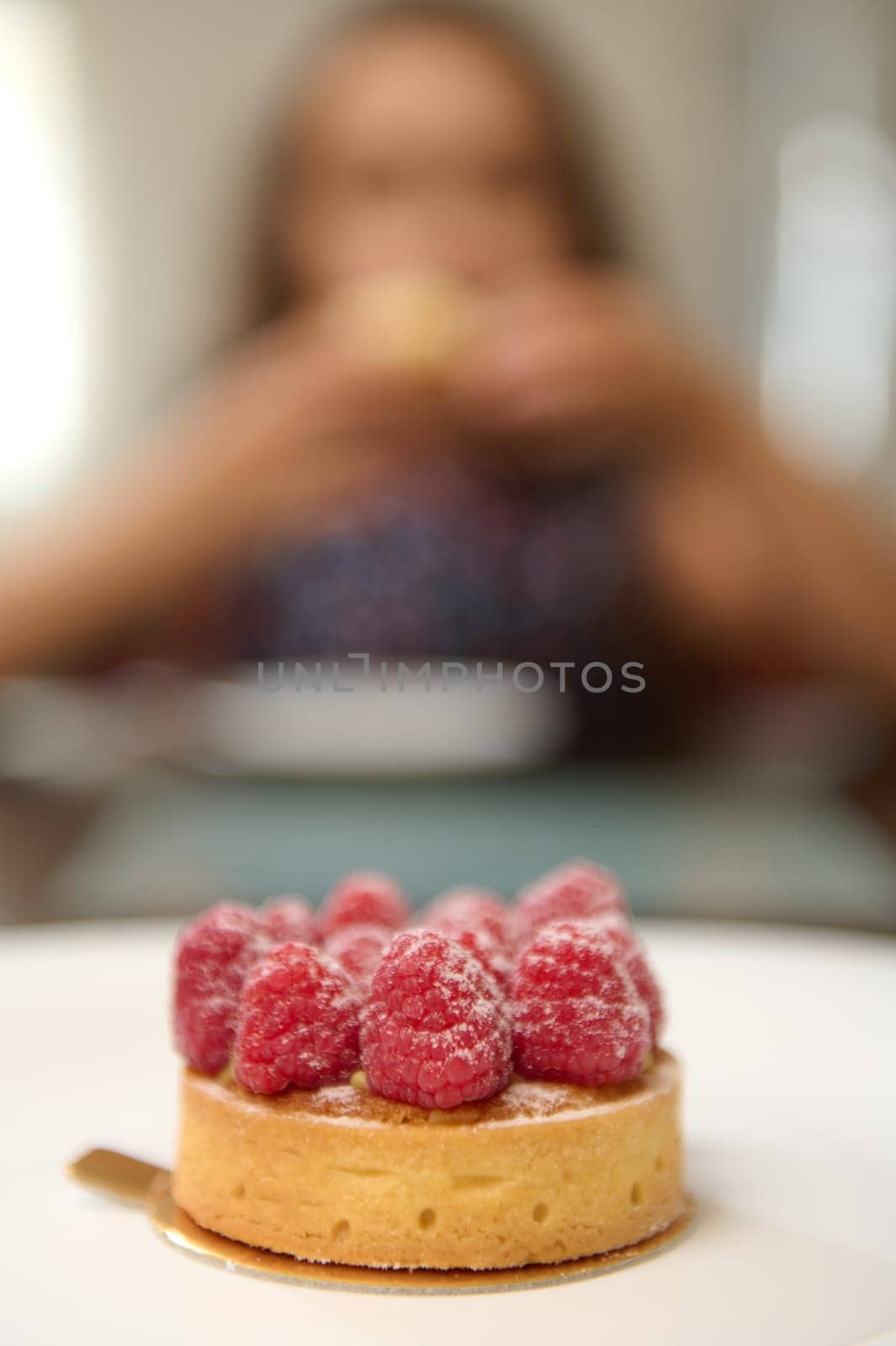 Details on a tasty sweet French dessert - a tartlet with raspberries on white plate, over blurred background of a little child taking her breakfast or snacking. Food still life. Culinary. Patisserie