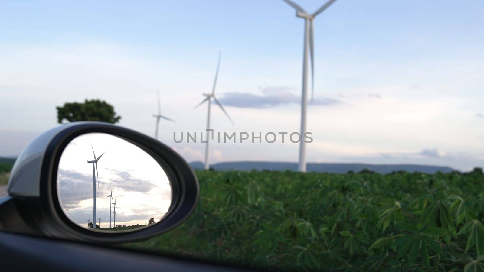 Progressive future energy infrastructure concept of wind turbine reflected in side mirror of electric vehicle being charged at charging station powered by green and renewable energy from wind turbine.