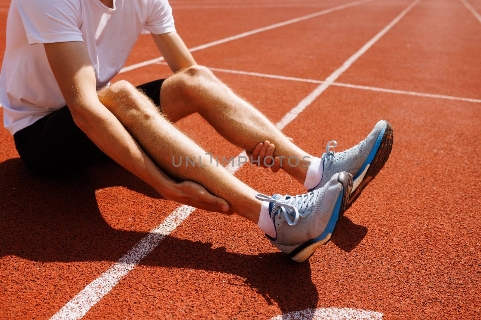 A runner sitting on a track holding his leg in pain, possibly due to a cramp or injury.