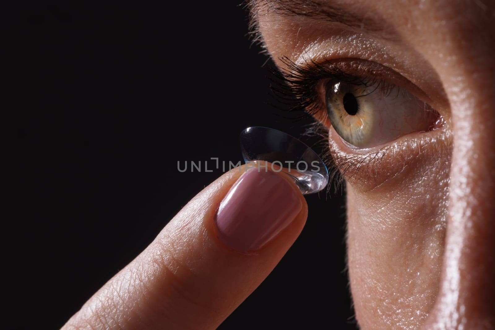 A close-up of a woman applying a contact lens to her eye. The focus is on her eye and the lens, showcasing the delicate process of lens application. The background is dark, emphasizing the subject.