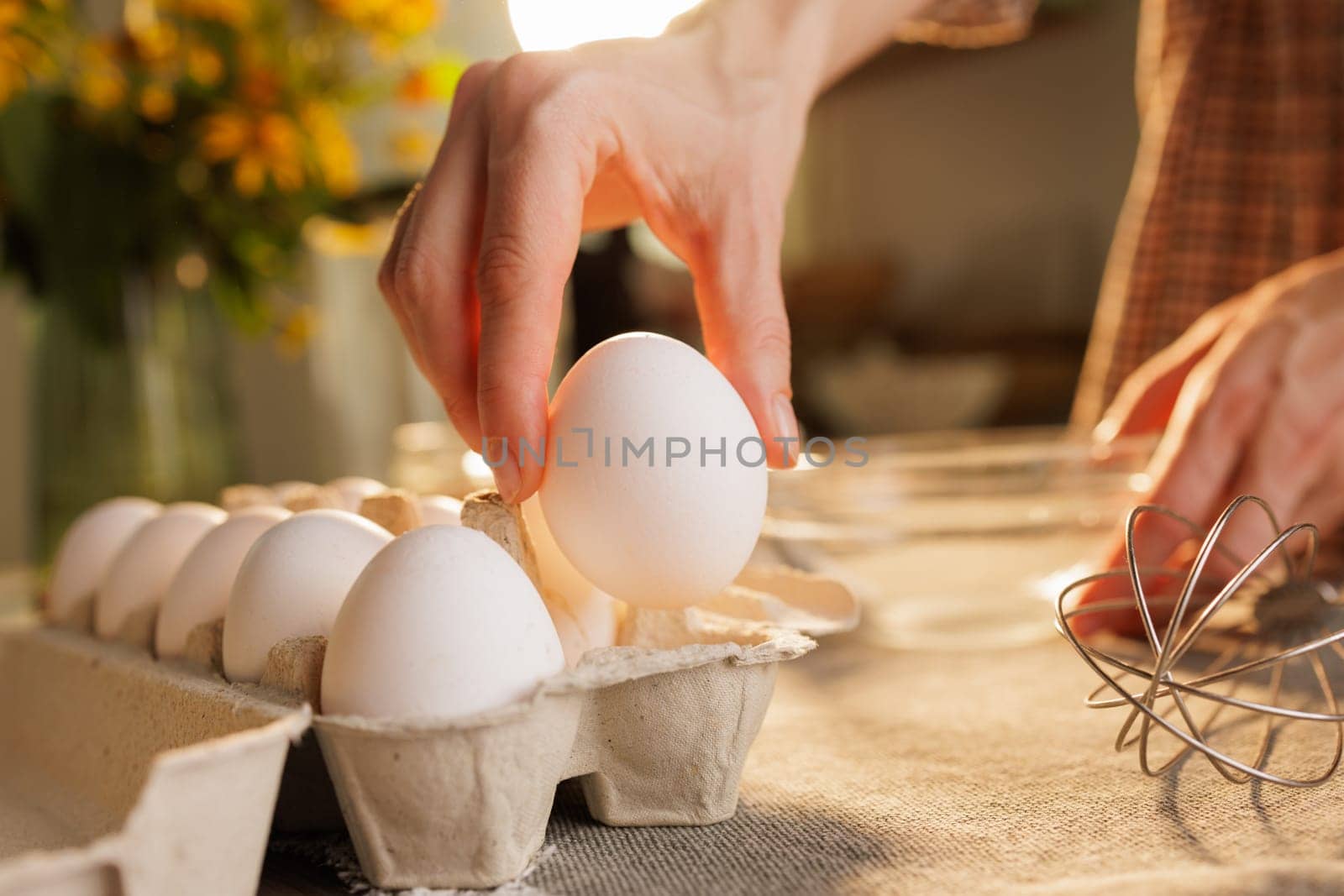 A hand carefully picks up a white egg from a carton, with a blurred background featuring a bouquet of flowers and kitchen utensils