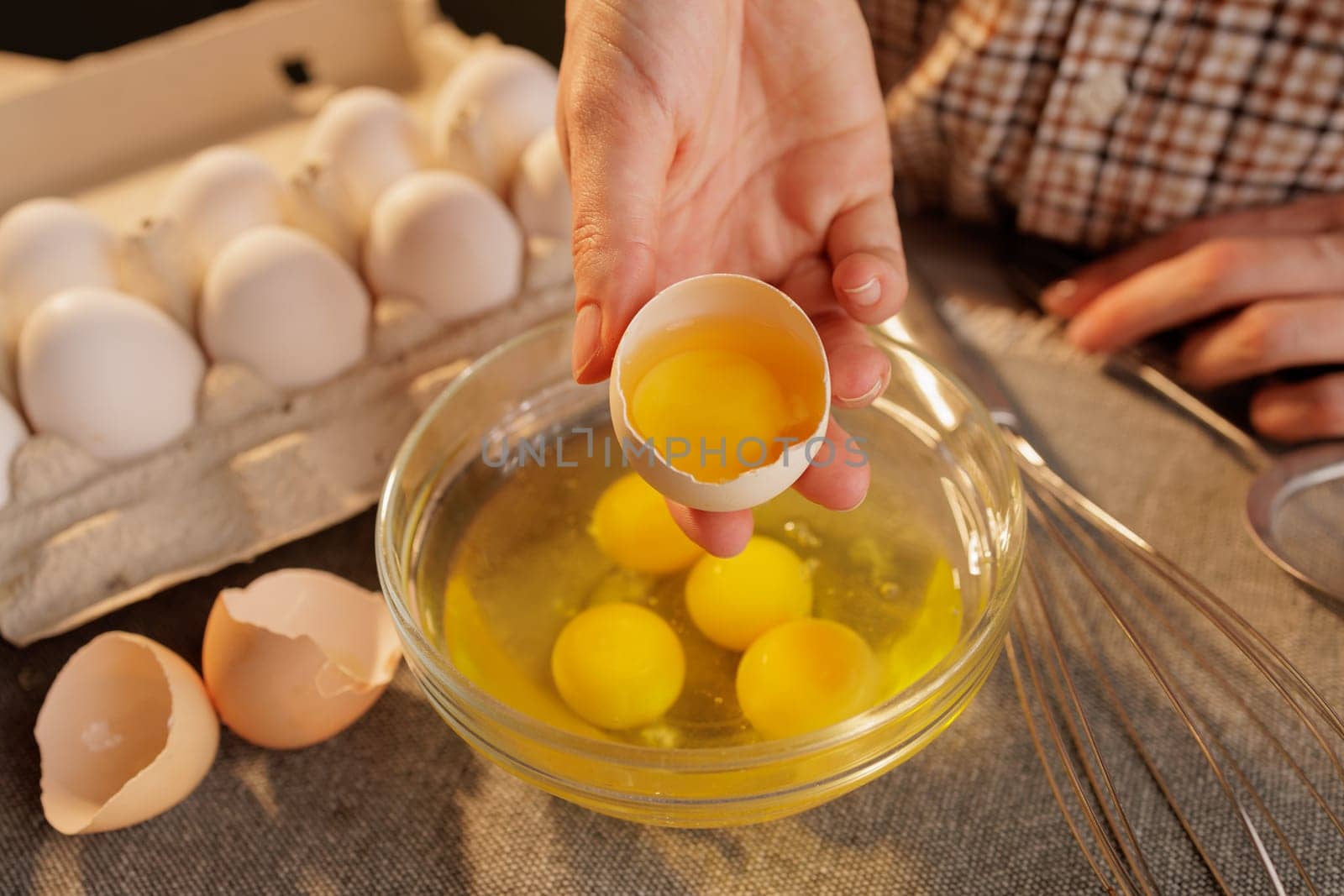A person cracking an egg into a glass bowl, with several eggs in an egg carton in the background. The scene is set in a kitchen, showcasing the process of preparing food.