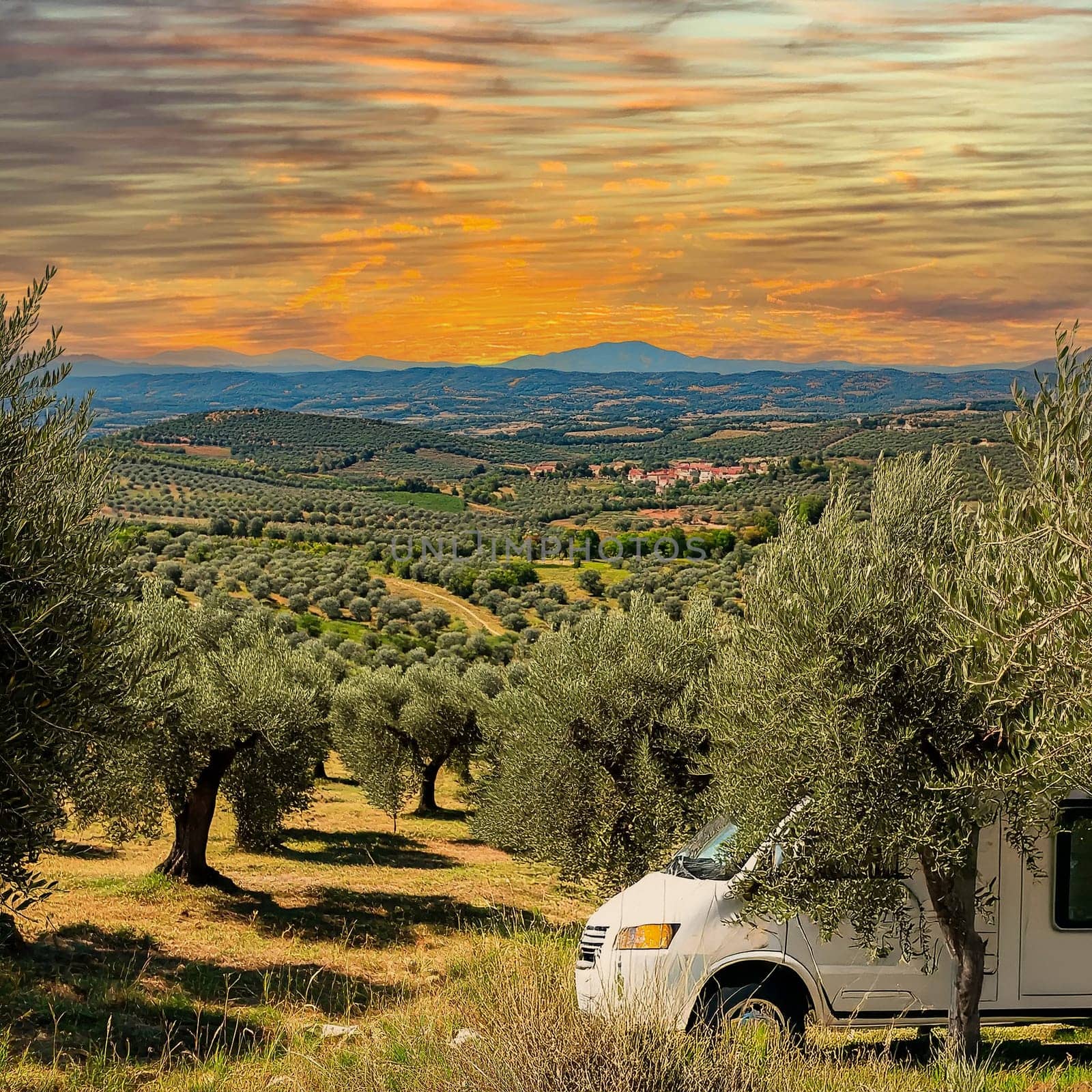 Camping car is parked on a green meadow surrounded by olive trees. The sun is just setting and is coloring the sky in warm orange and red tones.