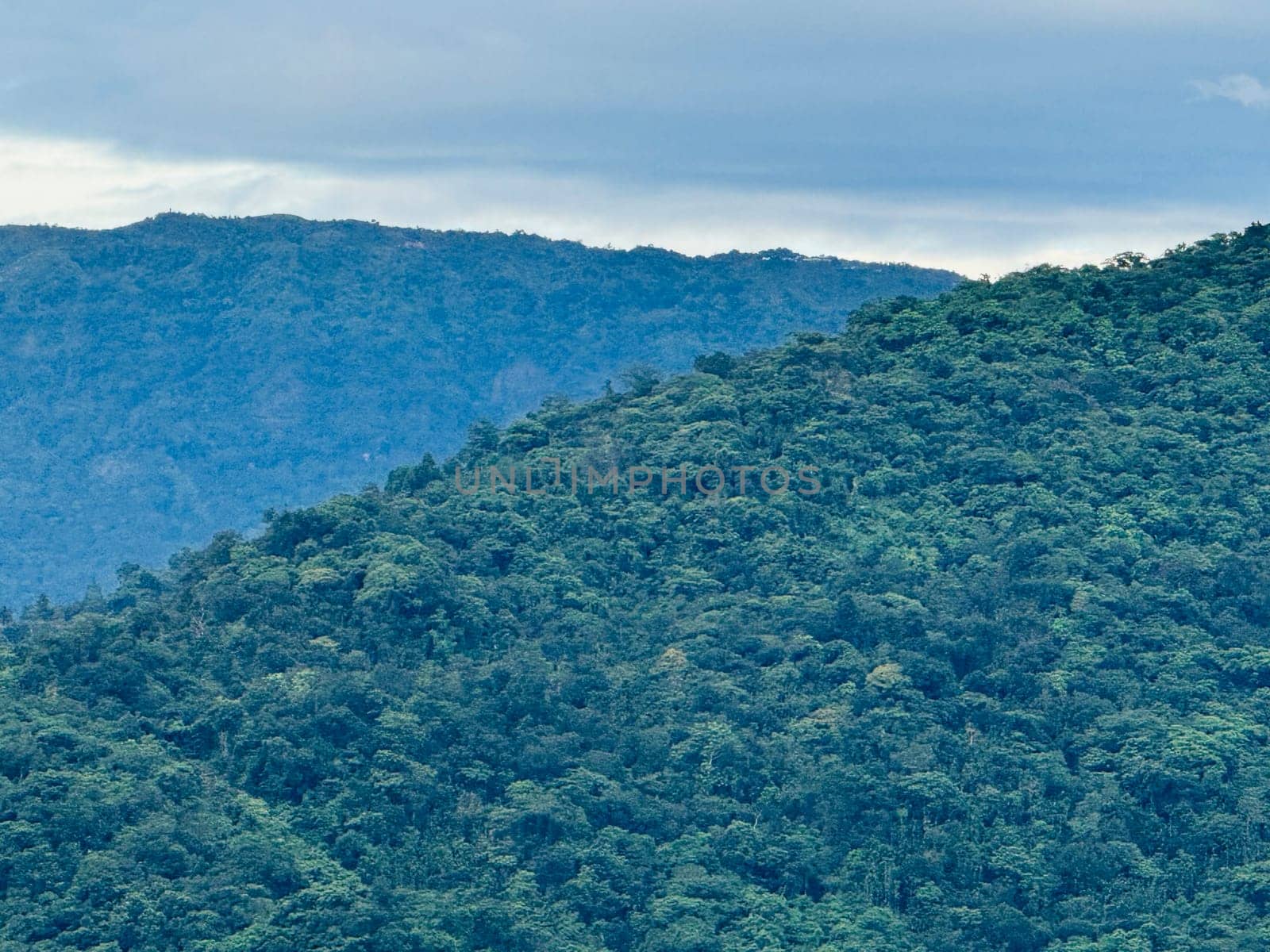 The vibrant green hills rise dramatically against a backdrop of a cloudy sky, revealing the dense vegetation and rich biodiversity typical of tropical rainforests.