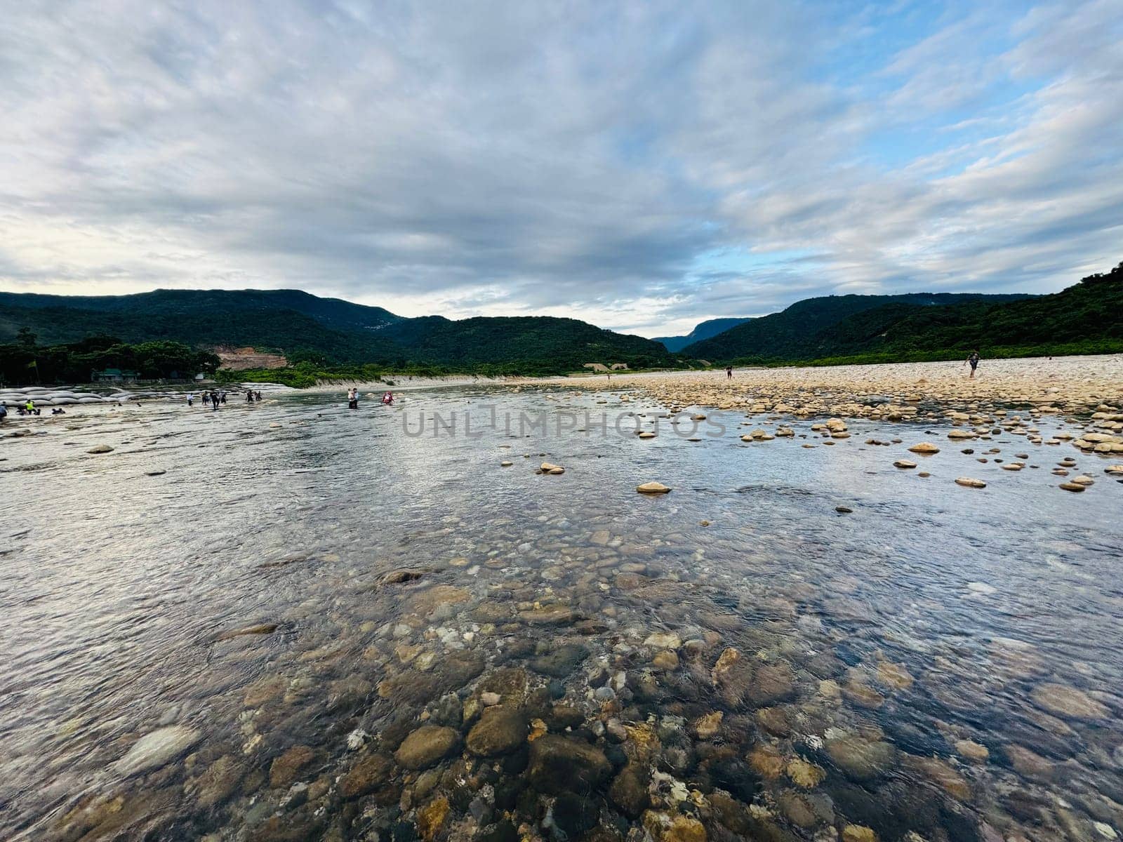 Visitors stroll along the clear waters of a riverbed, exploring smooth stones and enjoying the natural beauty of the surrounding hills under a partly cloudy sky.