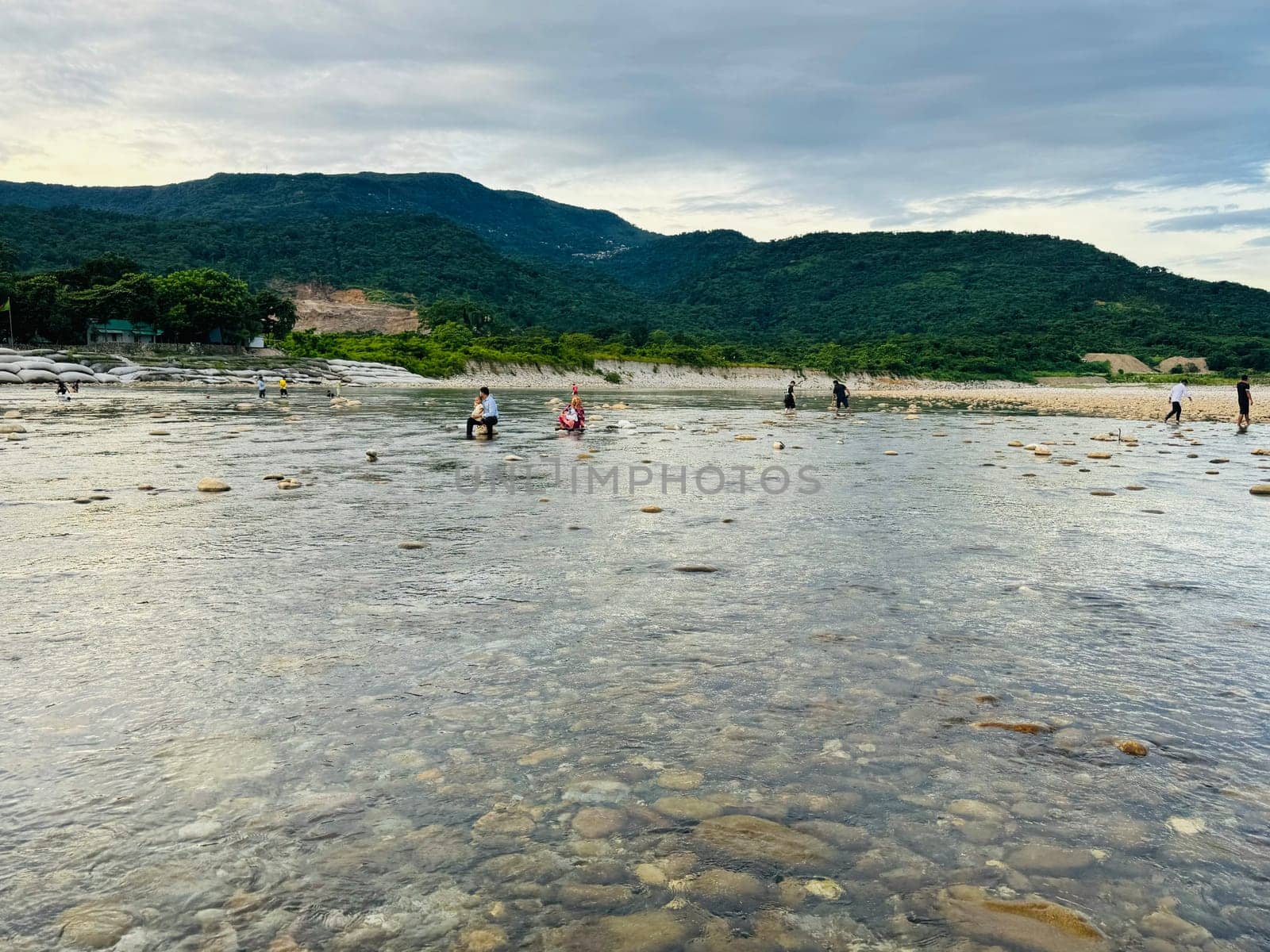 A group of individuals wades through shallow waters and rocks along the coastline, enjoying a tranquil afternoon in a picturesque natural setting with mountains in the background.