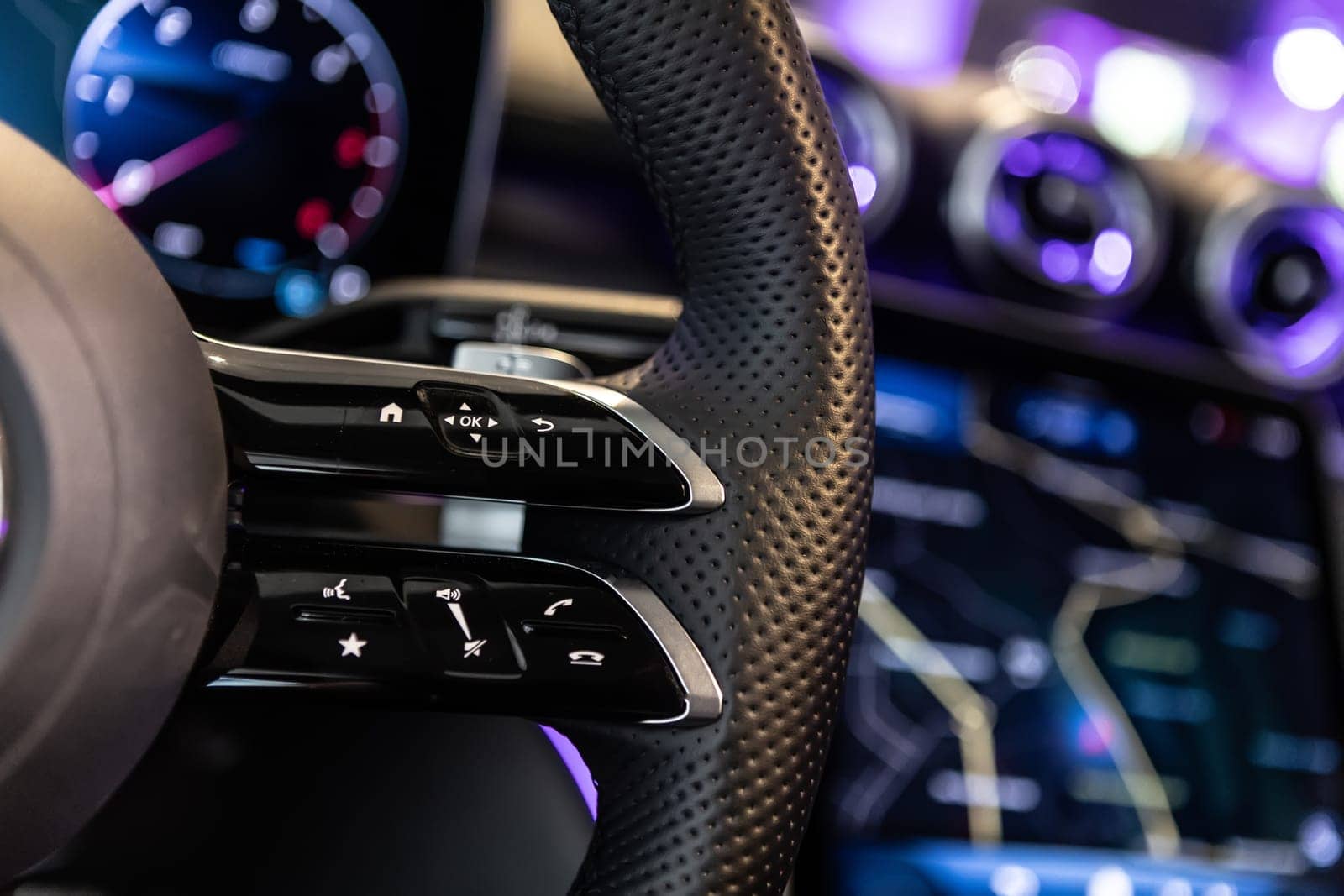 Close-up shot of a luxury car steering wheel featuring control buttons and sleek leather details, with a blurred background displaying a high-tech dashboard and navigation screen.