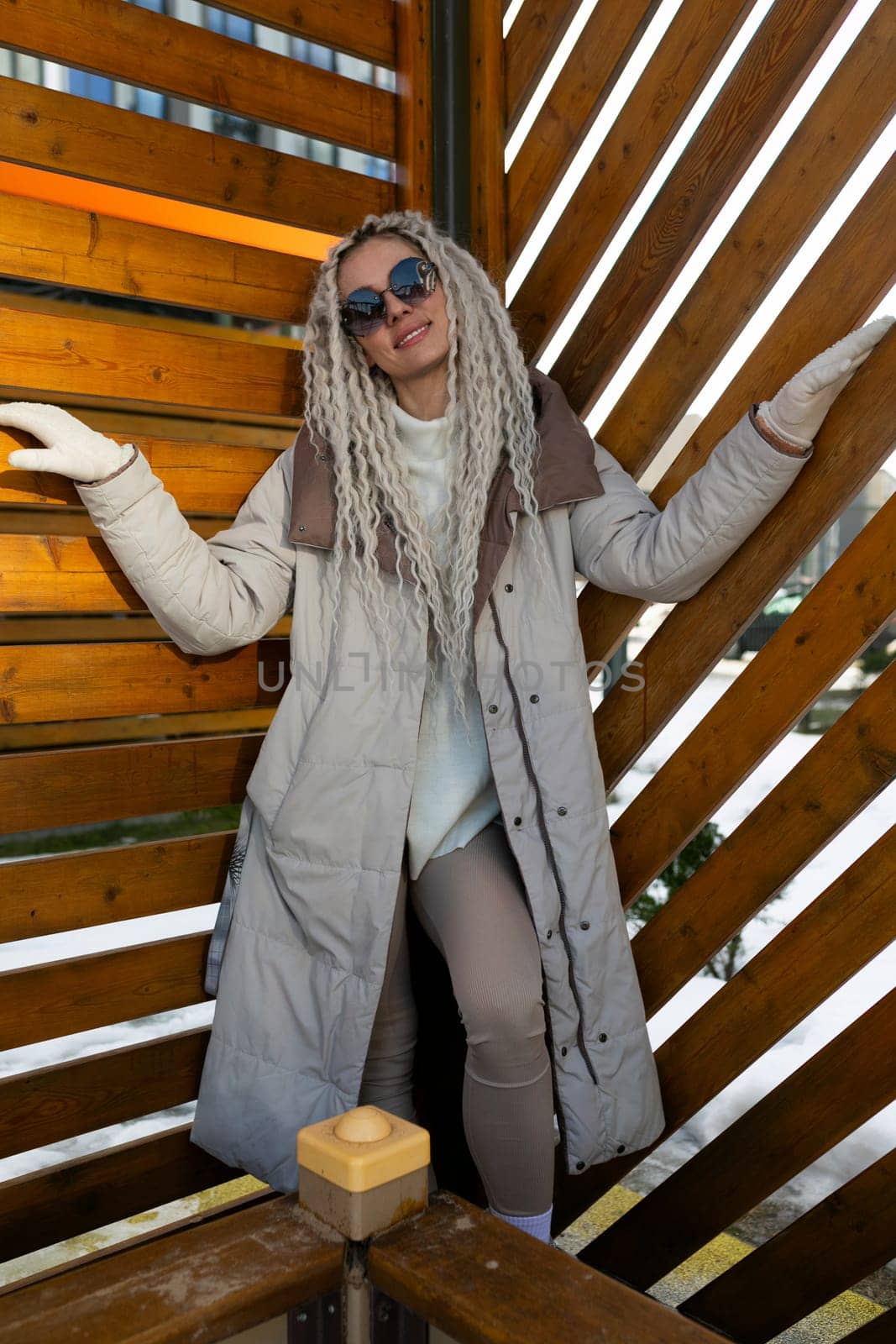 A woman with dreadlocks is standing in front of a wooden wall. She appears confident and relaxed, looking directly at the camera. The wooden wall behind her provides a rustic backdrop for the scene.