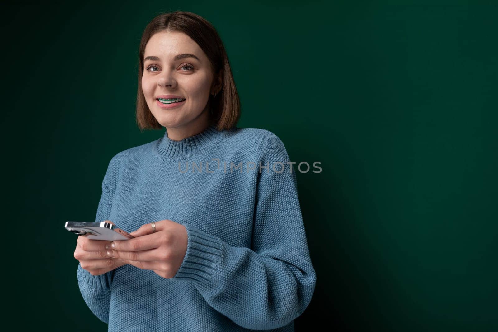 A woman wearing a blue sweater is seen holding a cell phone in her hand. She appears to be using the phone for communication or browsing. The background is simple and non-distracting.