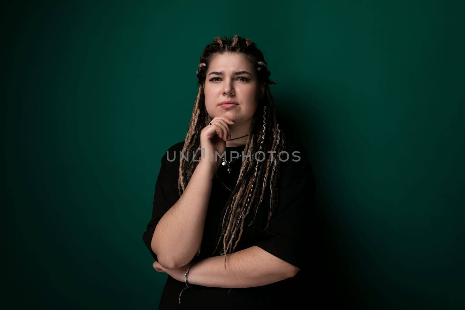 A woman with dreadlocks is leaning against a vibrant green wall. She appears relaxed and casual, with a confident posture. The contrast between her hair and the wall adds visual interest to the scene.
