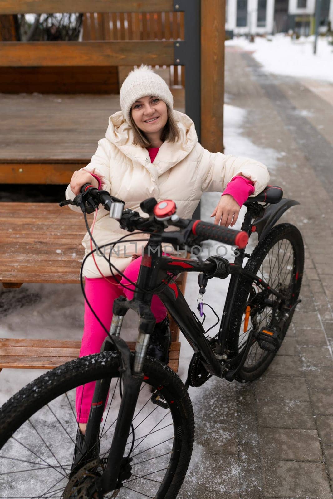 A woman is sitting on a bench next to a bike, with buildings and trees in the background. She appears relaxed and is looking at her phone.