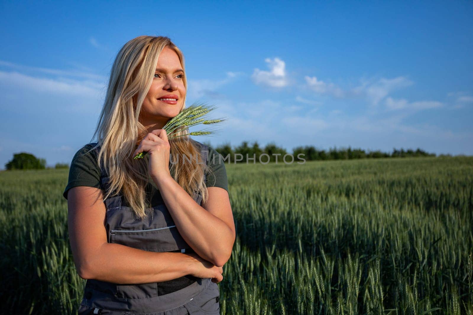 In the midst of a vast grain field, a content and smiling female farmer stands, holding wheat stalks near her face. Her radiant smile showcases her teeth as she cradles the stalks against her cheek, holding them in one hand. With a gaze fixed on the horizon, she exudes a sense of satisfaction and fulfillment. The golden sunlight bathes the scene, illuminating her features and adding a warm glow to the surroundings. Against the backdrop of the sprawling landscape, her presence exudes tranquility and harmony with nature. This captivating image captures the essence of agricultural contentment, where the beauty of the countryside meets the joy of a successful harvest. It celebrates the bond between farmer and land, where hard work and dedication yield abundant rewards, and each moment is cherished amidst the golden fields of grain