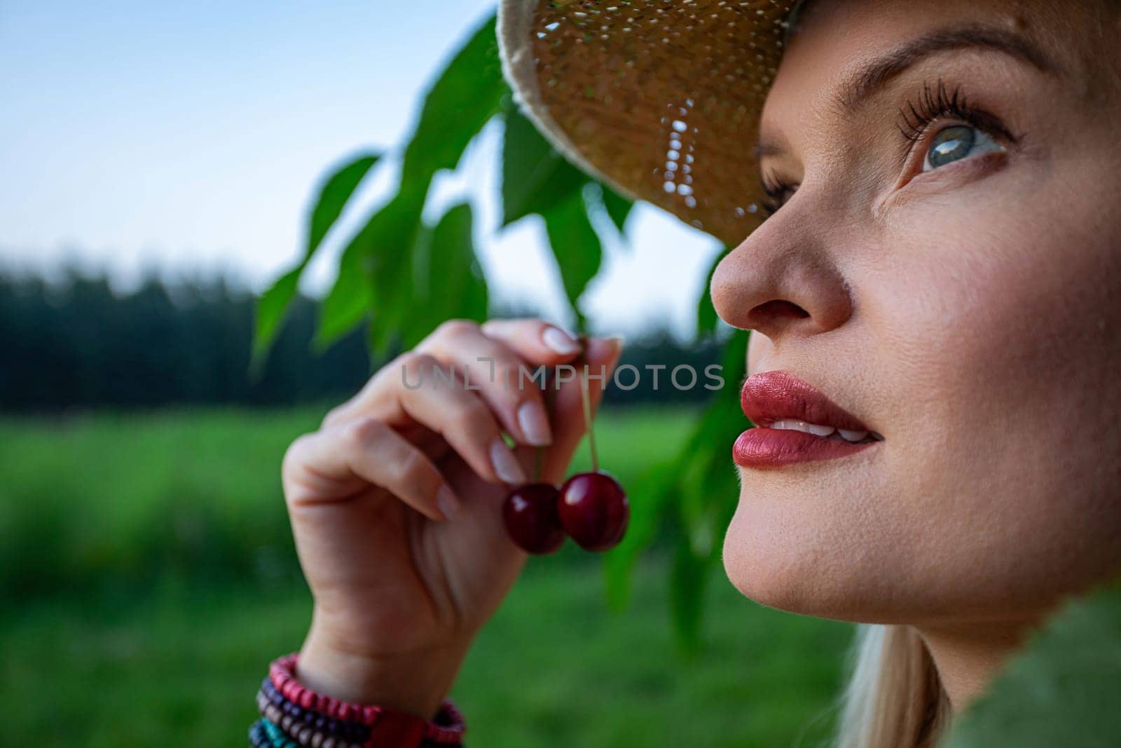 The close-up image captures the essence of a woman's face and hands, revealing her painted, full lips. She is holding red cherries in her hand. Her gaze is directed towards the sky, framed by a backdrop of a cherry tree branch and a blurred forest in the distance. Imperfections adorn her skin, adding a raw authenticity to her presence, while her piercing blue eyes reflect a sense of introspection. Against her wrist, a colorful bracelet crafted from beads adds a playful touch to her demeanor. She has straw hat on her head. The cherries in her grasp exude ripeness, promising a burst of sweetness with every bite. Amidst the tranquil surroundings of the orchard, she stands as a symbol of connection to nature, her contemplative gaze hinting at a moment of quiet reflection amidst the beauty of the natural world