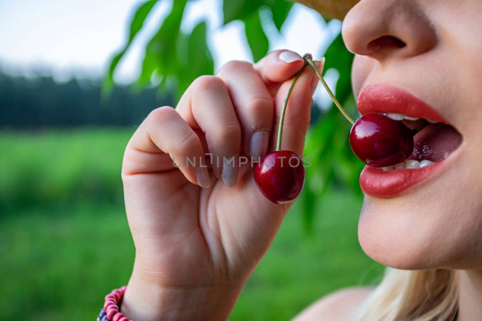 In the close-up image, a woman's face and hands take center stage, her lips painted and full, holding a single red cherry between her teeth. The cherry stem protrudes from the fruit, adding a touch of whimsy to the scene. Her white teeth and tongue are visible as she holds the cherry securely. In the background, a cherry tree branch is faintly visible, along with a blurred forest in the distance, creating a serene backdrop. The woman's expression is playful yet relaxed, her eyes twinkling with delight. The cherries she holds are vibrant red and ripe, ready to be savored. Against the soft focus of the forest, the details of the cherry and the woman's features are highlighted, drawing the viewer's attention to the simple yet captivating moment captured in the image