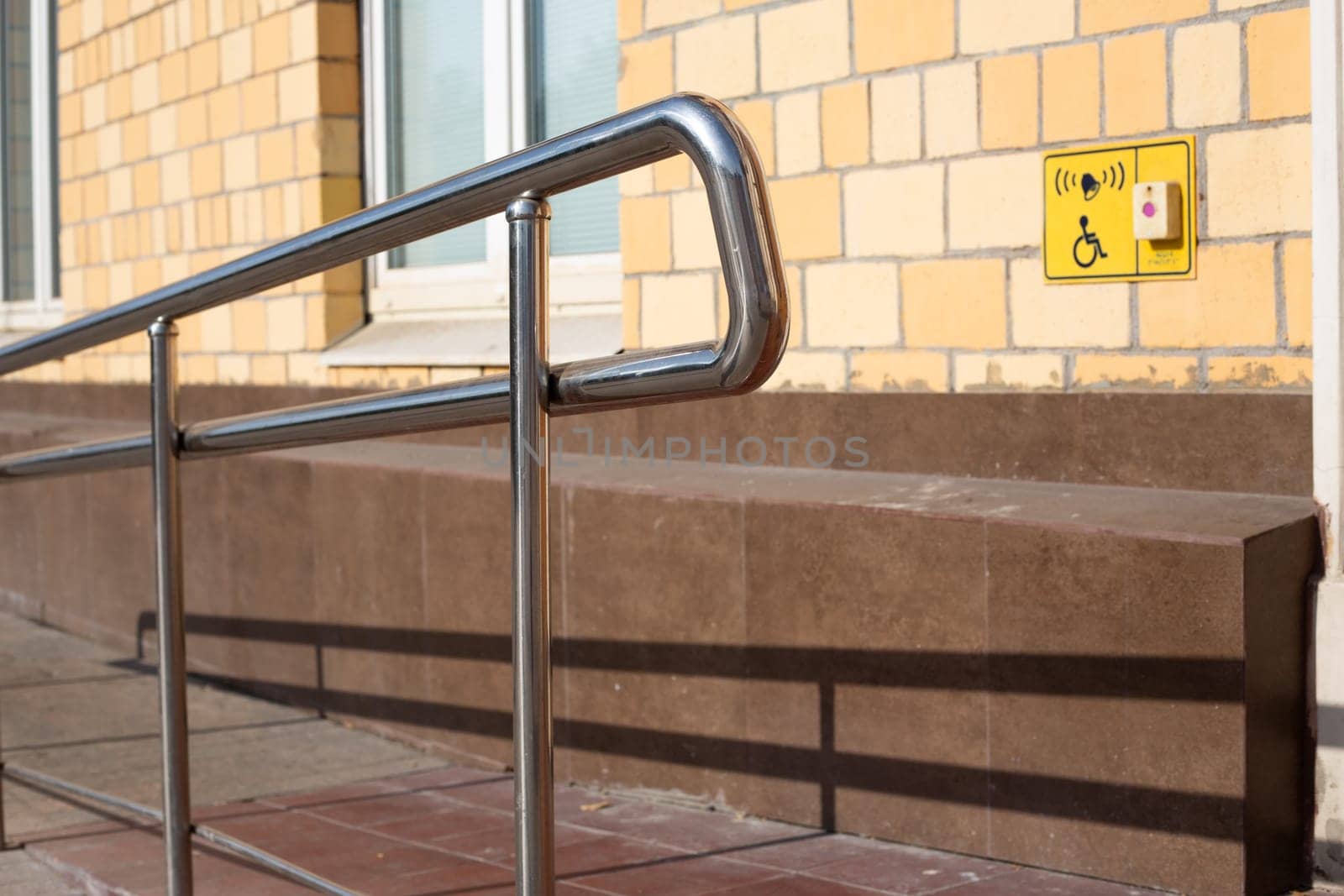 Metal railing is next to yellow sign that says handicap accessible with help button. Ramp for people in wheelchairs at the entrance to building with handrails for assistance