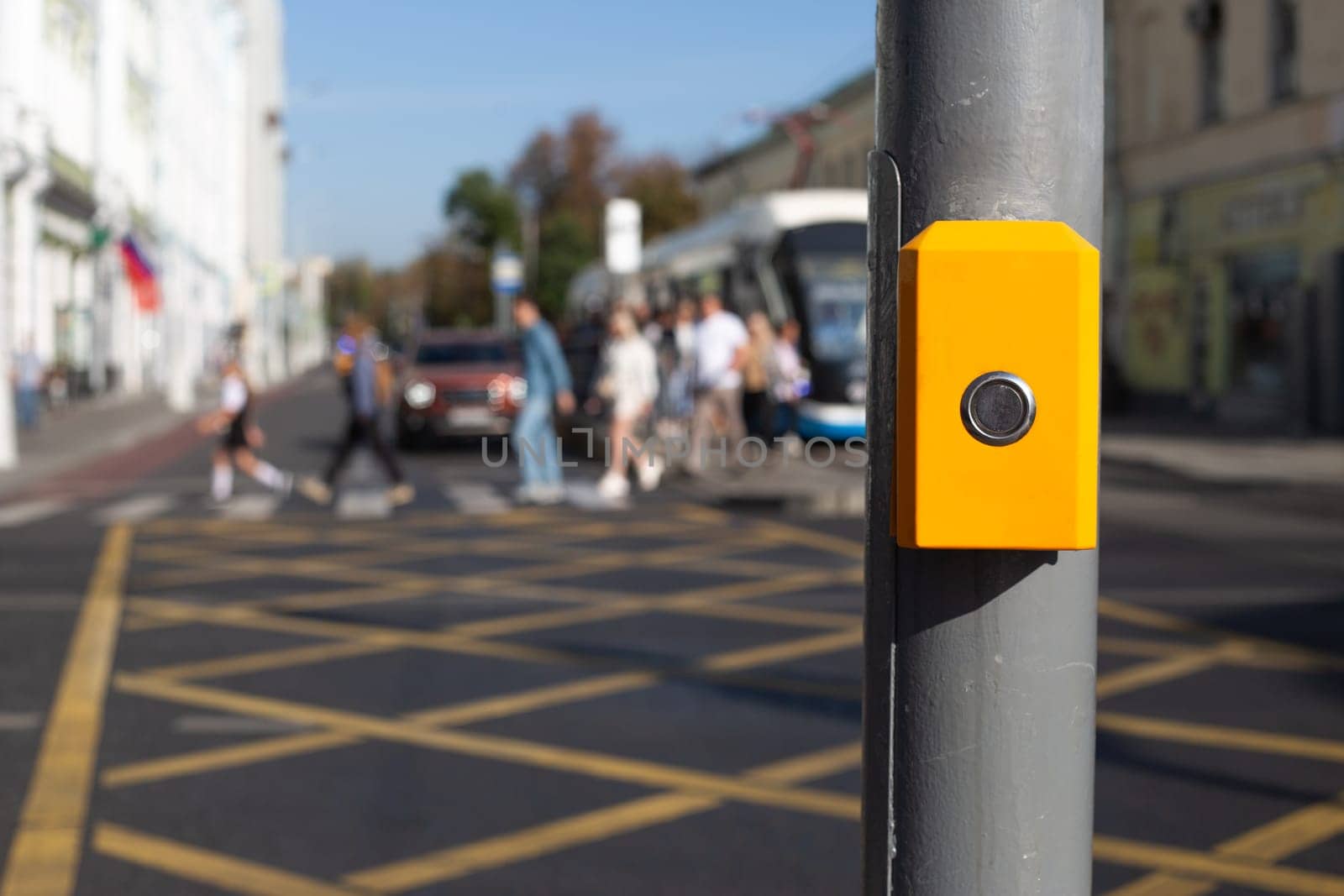 Button for changing traffic light signal for pedestrians to cross an intersection. Control panel for waiting to cross the road hangs on pole on the background of people cross road
