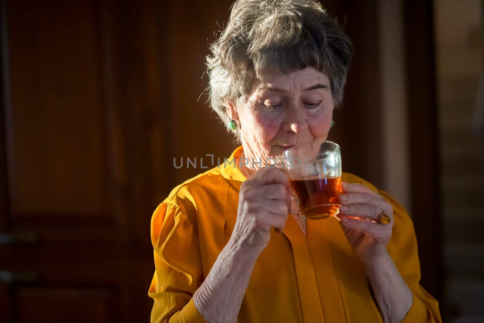 An elderly well-groomed gray-haired woman holds a glass cup of tea in her hands and enjoys its taste, the grandmother has a good time in her cozy house, relaxing in the kitchen in the rays of the sun.