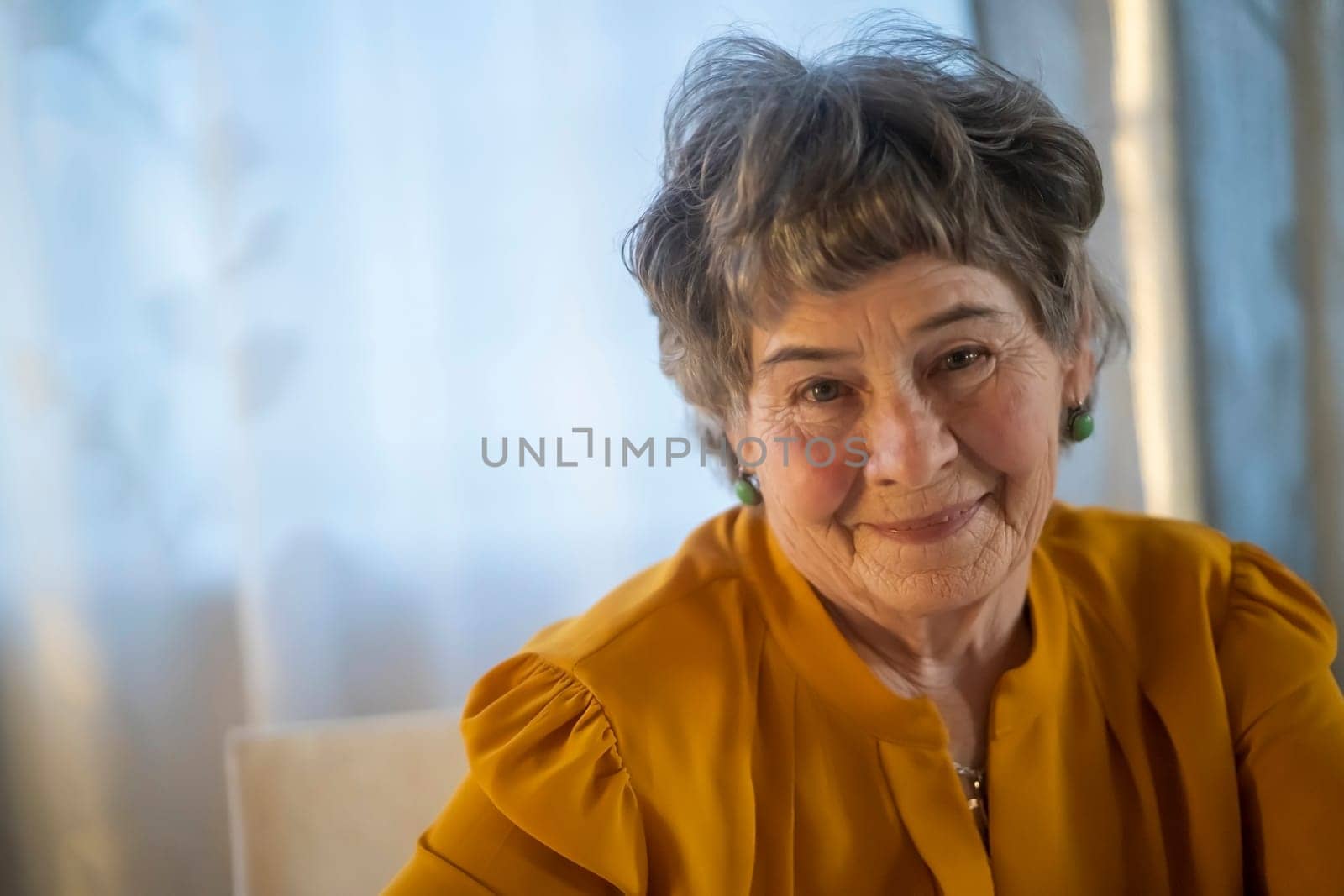 A portrait of a laughing, smiling elderly woman with gray hair and wrinkles on her face, but young at heart and leading an active lifestyle. Happy pensioner is pretty posing in her cozy home.