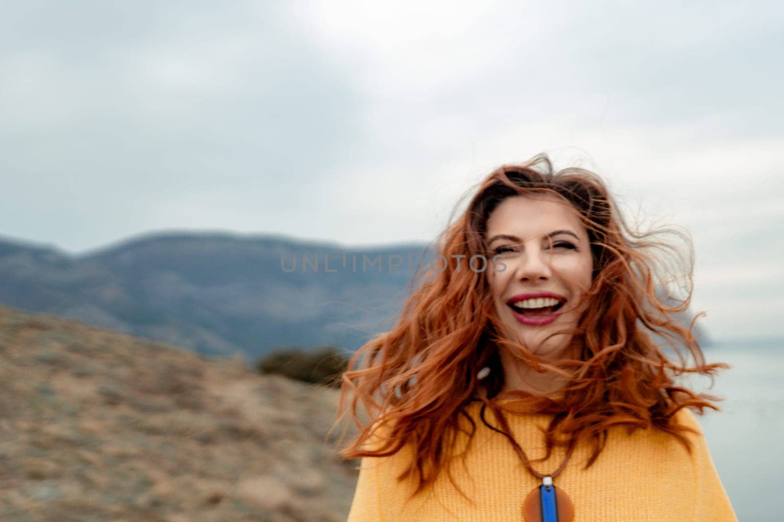 Portrait windswept hair happy woman against a backdrop of mountains and sea. Daylight illuminates the tranquil outdoor setting.