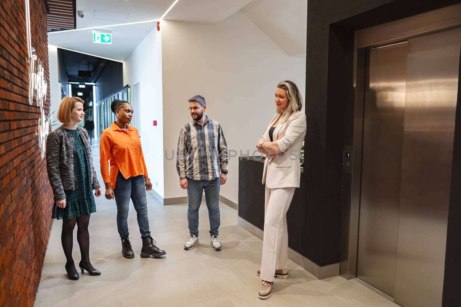 A diverse group of individuals of varying ages and genders are standing together in a hallway, appearing to engage in conversation or waiting for something. The hallway is well-lit and lined with doors on either side.