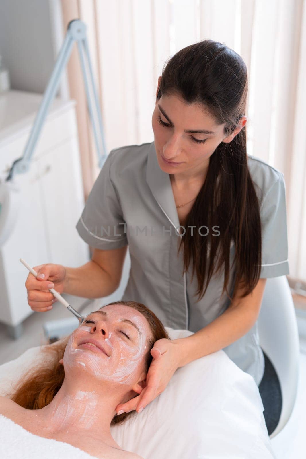 Professional cosmetology. Beautician at spa salon applying face peeling mask to young woman, relaxed female enjoying wellness and skin care treatments.