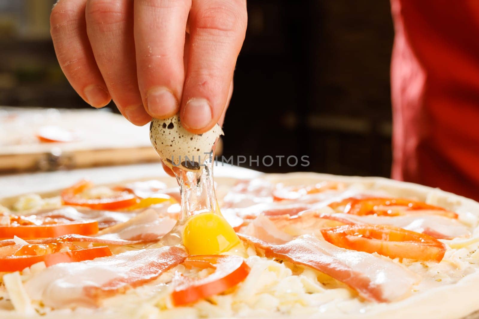 A close-up of a hand cracking a quail egg onto a pizza topped with ham, cheese, and sliced tomatoes. The pizza is uncooked, showcasing fresh ingredients and a rustic kitchen background.