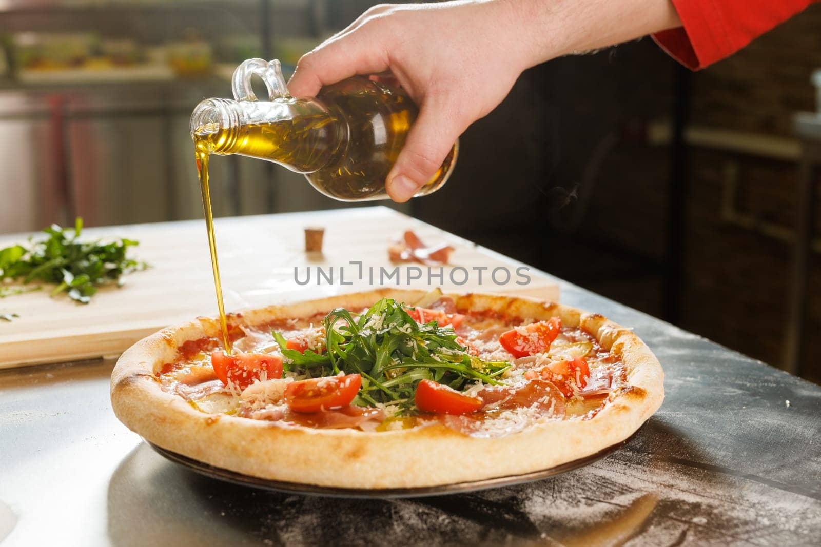 A hand pouring olive oil onto a freshly baked pizza topped with arugula and tomatoes in a kitchen setting.