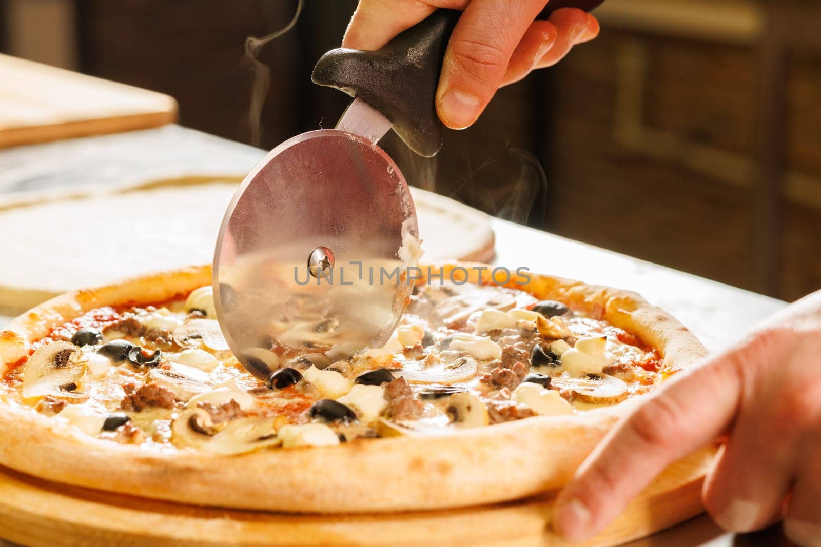 Close-up of a person cutting a freshly baked pizza with a pizza cutter. The pizza has toppings including mushrooms, olives, and cheese.