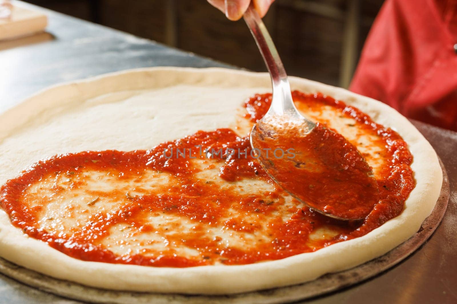 A person spreading tomato sauce on pizza dough with a ladle in a kitchen.