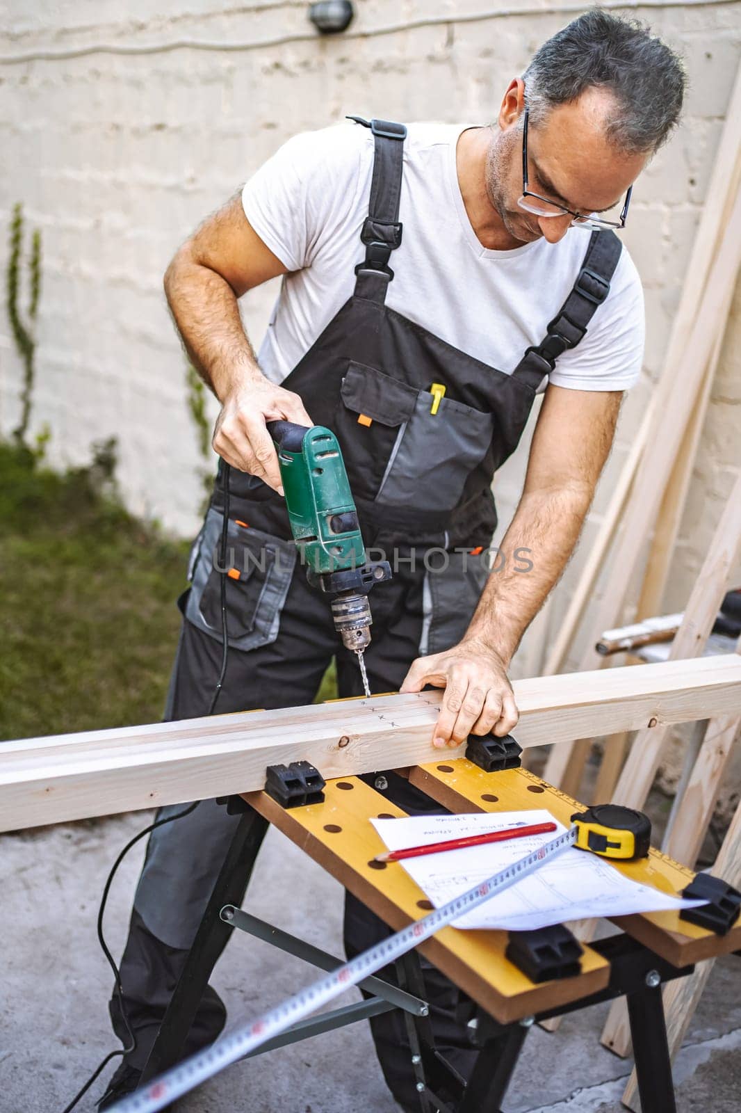 Mature male carpenter using electric screwdriver on a wooden plank outdoors.