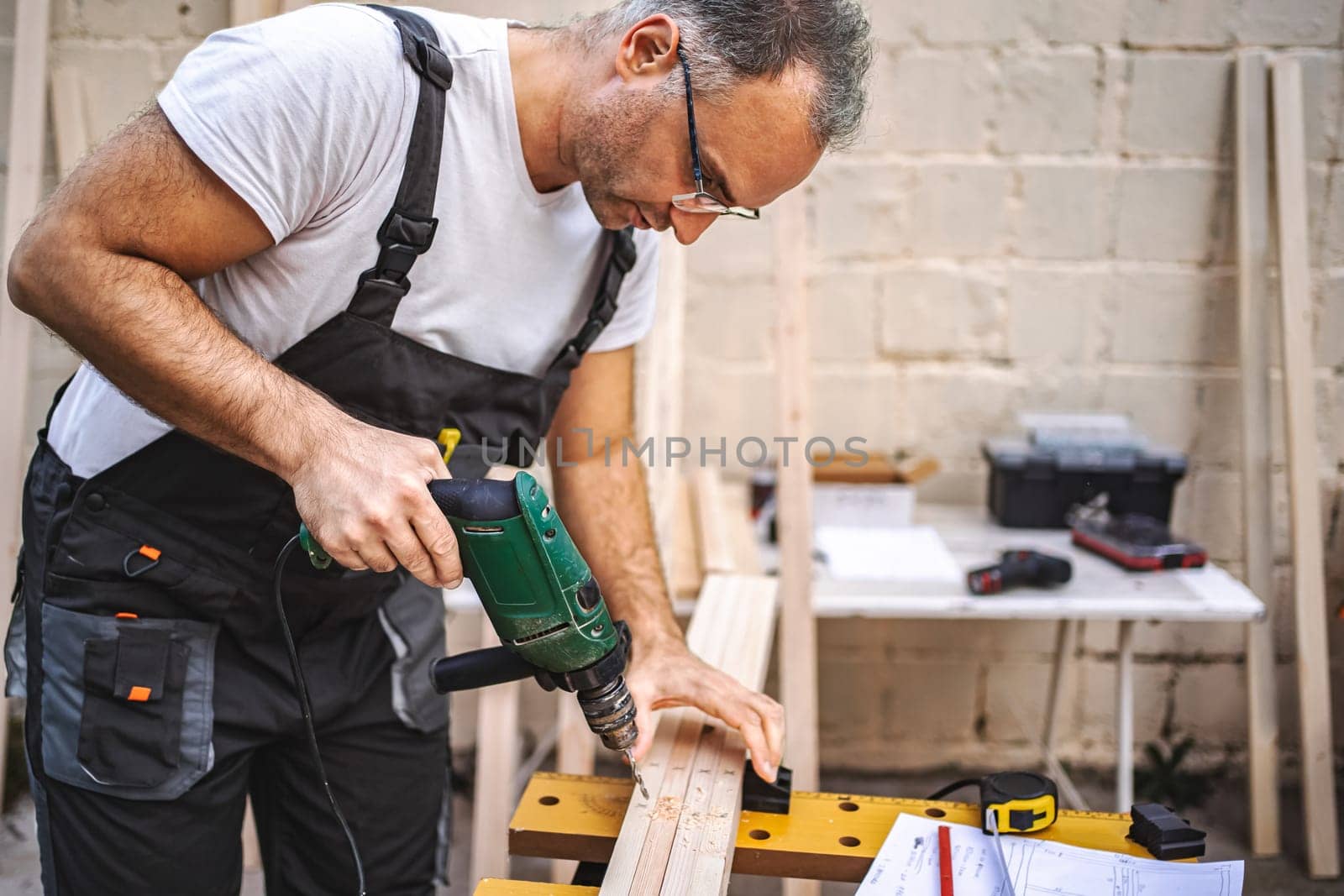 Mature male carpenter using electric screwdriver on a wooden plank outdoors.