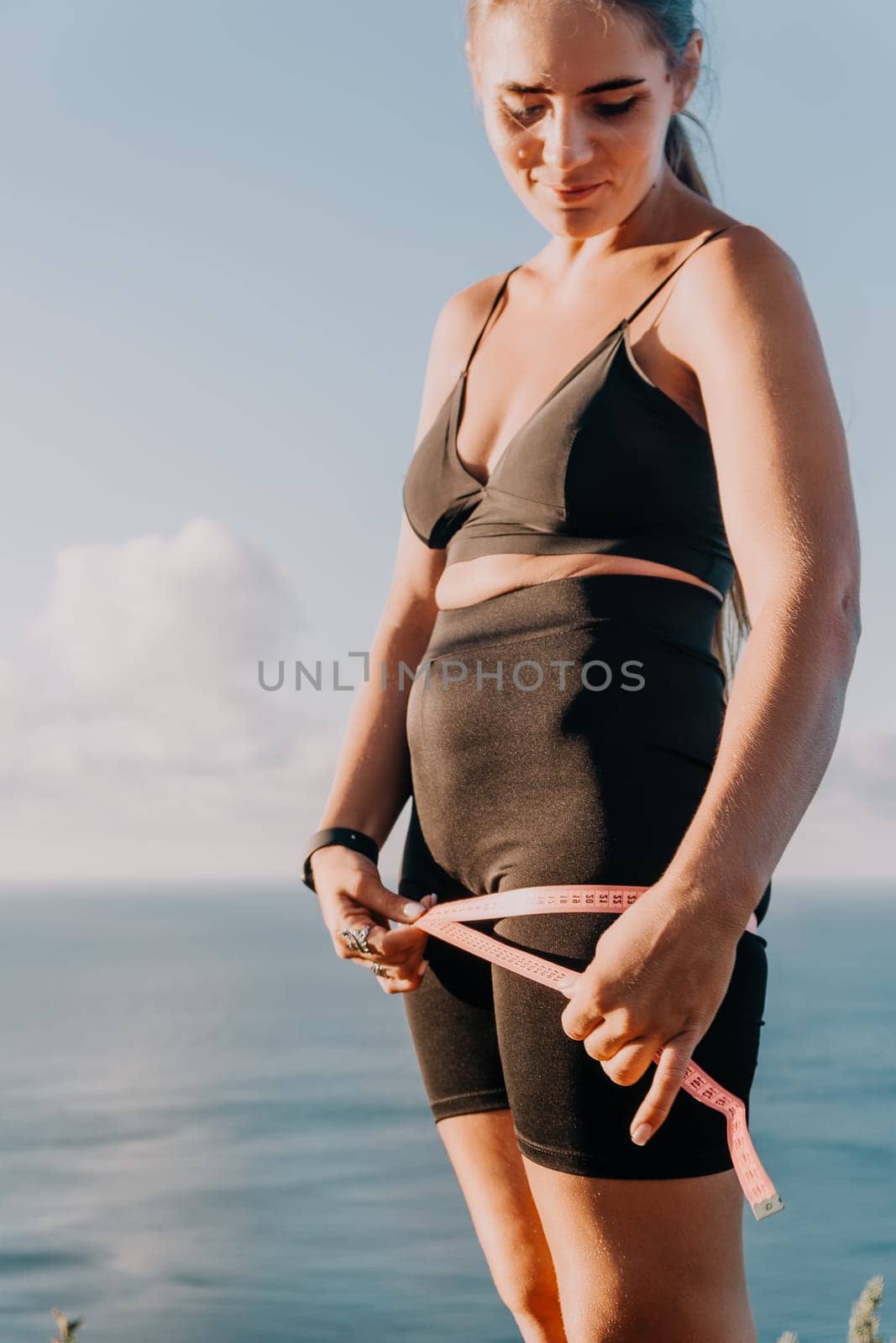A woman in athletic wear measures her waist with a tape measure while standing on a cliff overlooking the ocean.